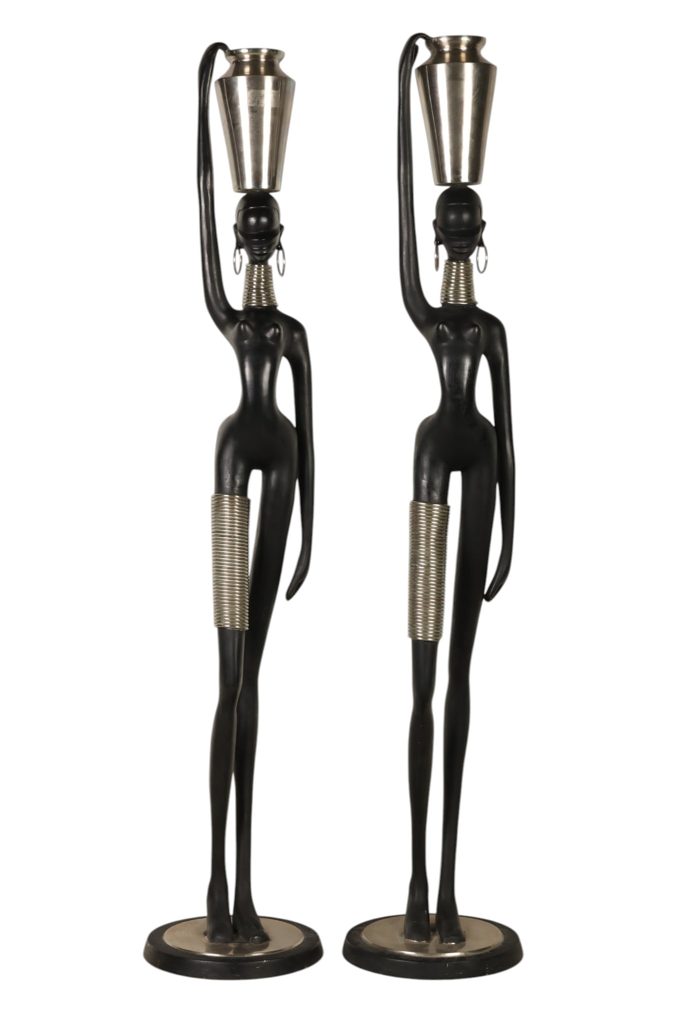 Pair of Hagenauer Style MCM Nubian Woman with Vase: Pair of Mid Century Modern sculptural 66 inch figures in the manner of Hagenauer, depicting elongated African women in stylized form. Each figure is rendered in a matte black finish with silver-tone c