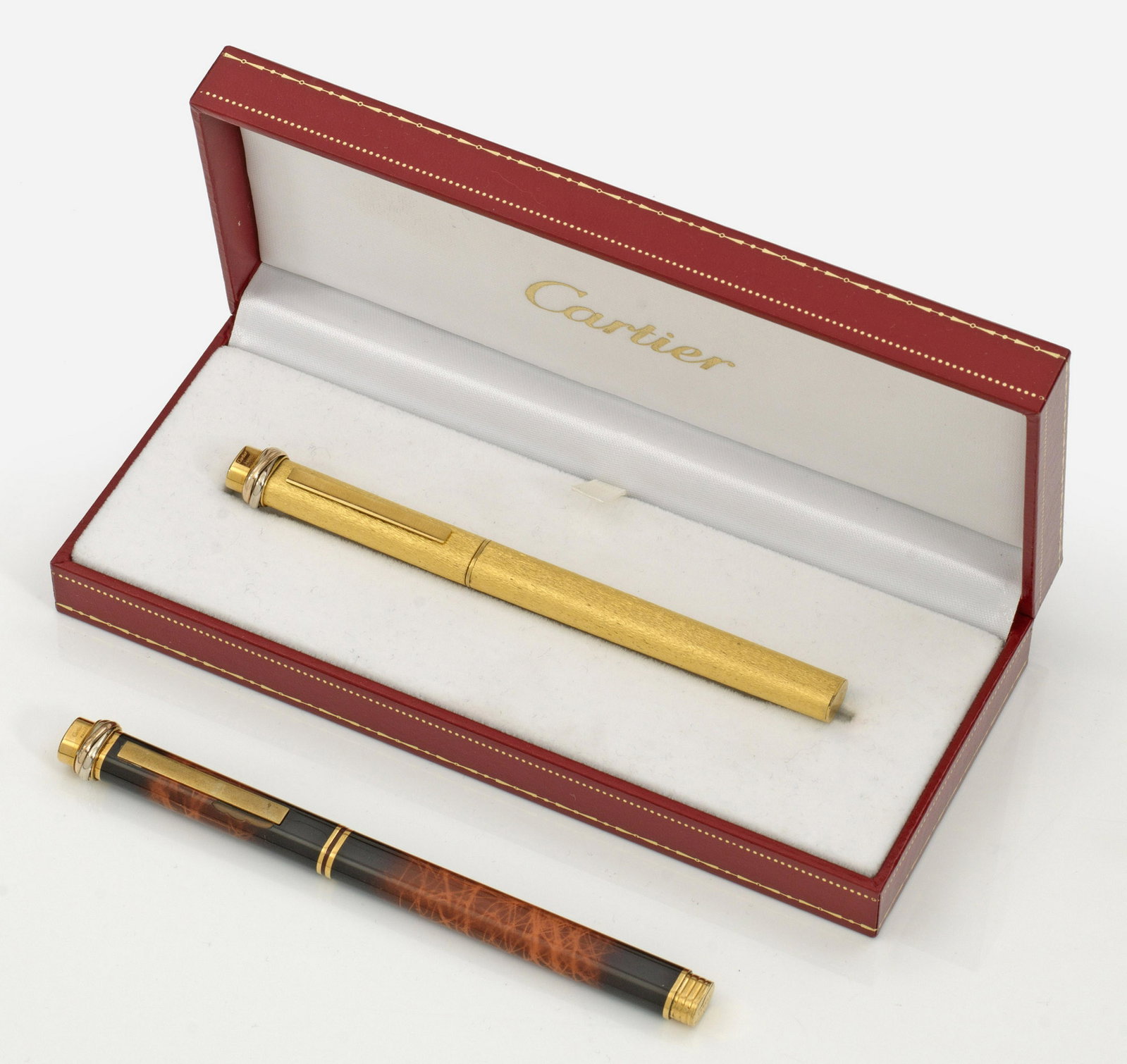 Zwei "Must de Cartier" Kugelschreiber: Metall, vergoldet. Fein ziselierter Korpus bzw. Chinalack in Schildpatoptik. Oberhalb eingelassener Trinity-Ring. Herstellerzeichen. Teilw. besch.; Dazu Cartier Box. Vgl. Clais, Les must de Cartier. S