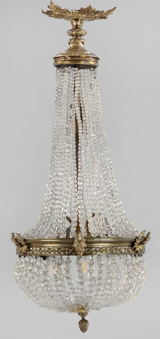Louis XVI-Deckenlüster: Messing und farbloses Glas. Balusterförmiger Korpus aus Prismenschnüren im Verlauf. Schmaler Reif mit reliefierten Lorbeerranken sowie oberer Abschluss in Form einer großen Blattkartusc