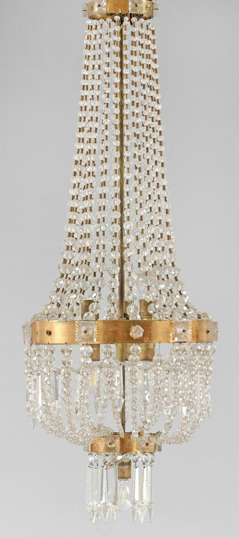 Großer Deckenlüster im Empire-Stil: Kupfer, Messing und farbloses Glas. Balusterförmiger Korpus aus Prismenschnüren im Verlauf. Unterer Abschluss aus großen, facettierten Prismenstäben. Innen sieben Lampenfassungen. 