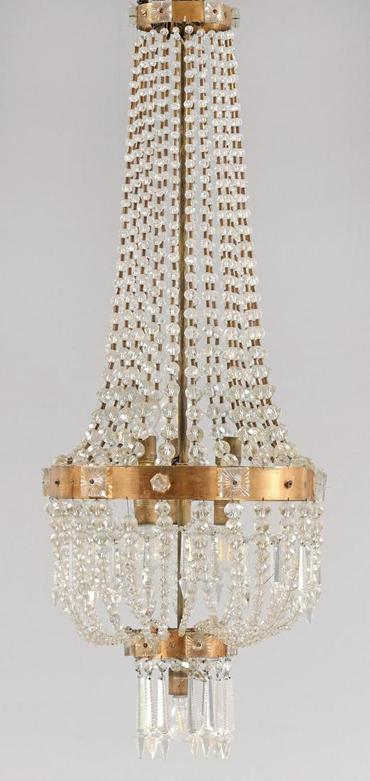 Großer Deckenlüster im Empire-Stil: Kupfer, Messing und farbloses Glas. Balusterförmiger Korpus aus Prismenschnüren im Verlauf. Unterer Abschluss aus großen, facettierten Prismenstäben. Innen sieben Lampenfassungen. 