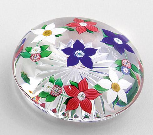 Paperweight "Corona" von Saint-Louis: aus limitierter Edition. Farbloses Glas mit Bodenschliffrosette. Gedrückte Form. Eingeschmolzene Girlande aus roten, weißen und blauen Clematis und kleinen Millefiori-Blumen umgibt eine zent