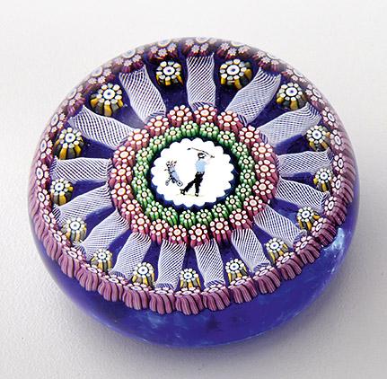 Paperweight mit Millefiori und Golfer-Silhouette: von Perthshire Paperweights Farbloses Glas. Halbrund. Auf dunkelblauem Grund zentrales Medaillon mit der Darstellung eines Golfers beim Abschlag, umgeben von Kränzen aus Millefiori-Canes und wei&