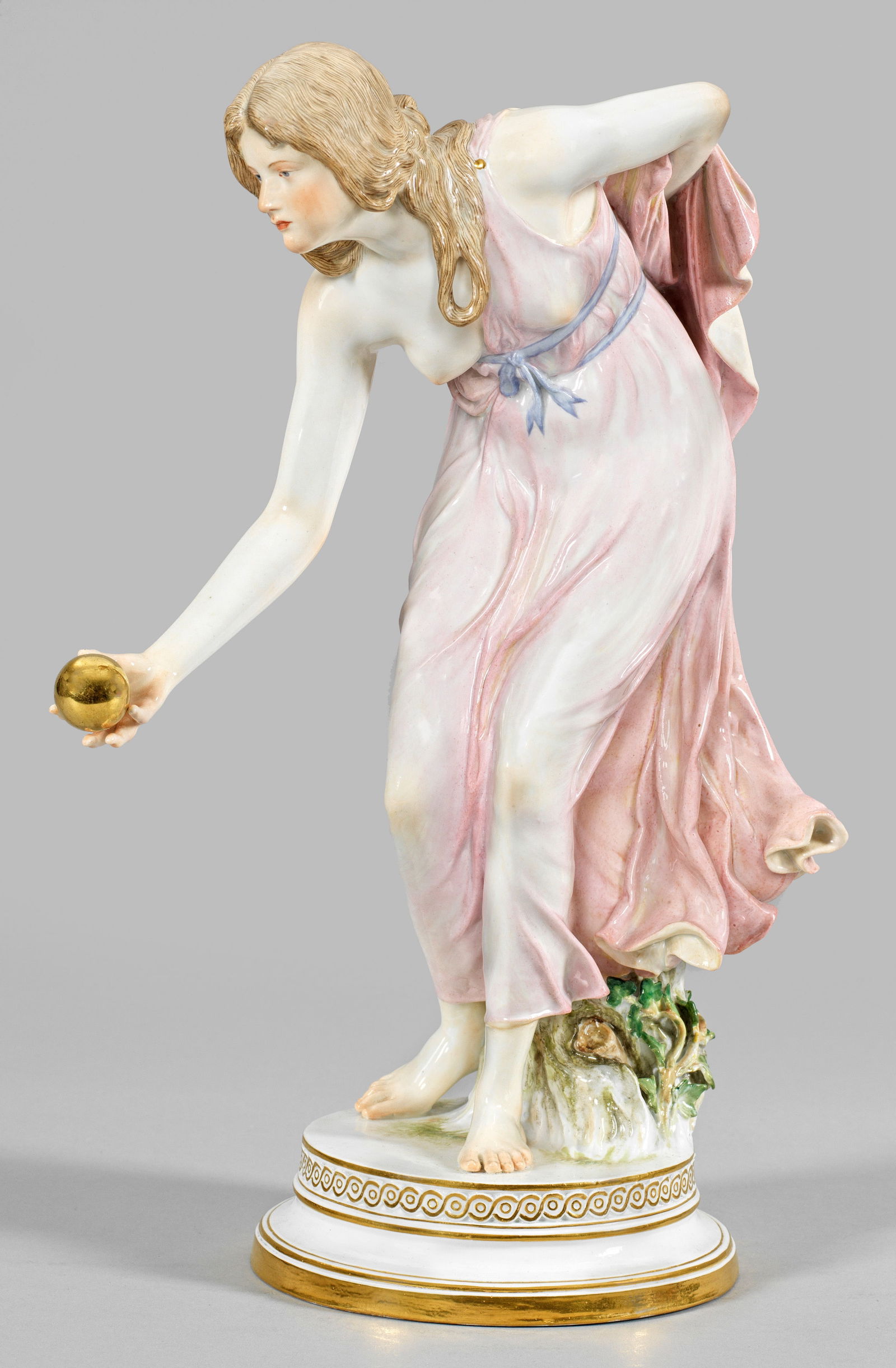 Jugendstil-Figur "Kugelspielerin" von Walter Schott: Auf rundem, profiliertem Sockel mit Flechtbandbordüre, stehendes, sich vorbeugendes Mädchen in einem hauchdünnen, den Körper umschmeichelndem, rosafarben staffiertem Kleid, in ihre