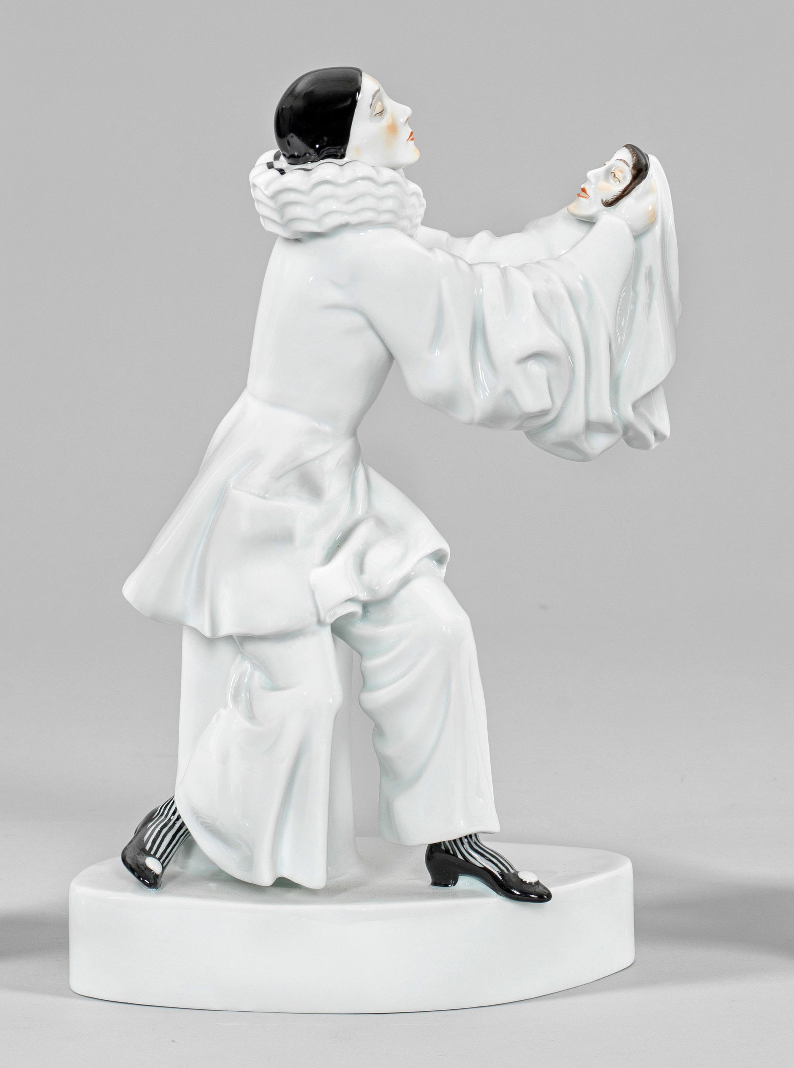 Bajazzo (oder Pierrot) mit Maske: Neuausformung eines Art Déco-Modells der Schwarzburger Werkstätten. Über linsenförmigem Sockel, auf einem Podest sitzender Bajazzo in langem Kostüm, eine Frauenmaske in seinen