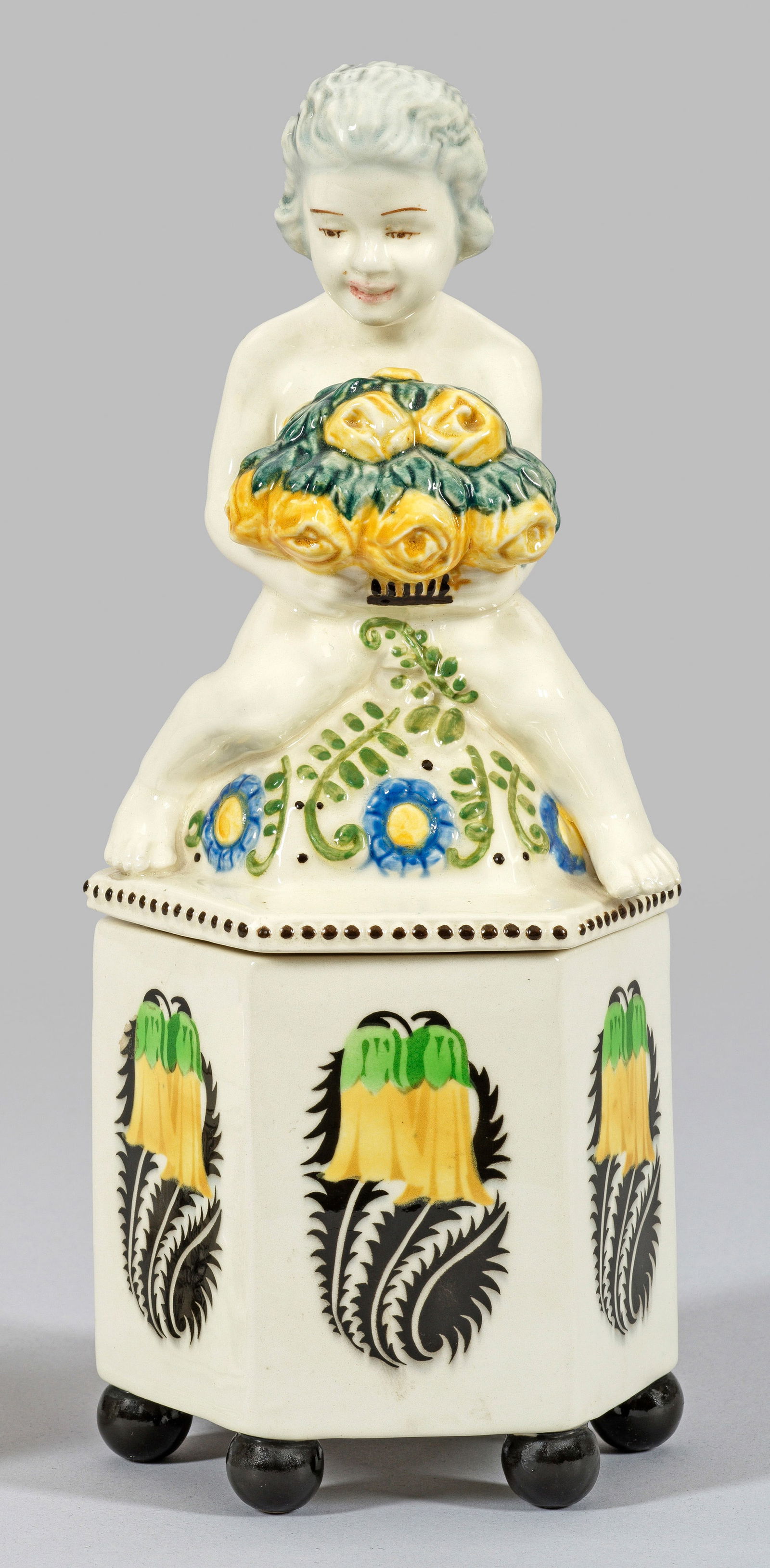 Zierdose mit Rosen tragendem Putto von Carl Klimt: Steingut. Sechseckig, Kugelfüße, reliefierter bzw. plastischer, glockenförmiger Deckel. Polychrome Unterglasurbemalung, auf dem Unterteil ein wiederkehrendes florales Motiv. Modell-Nr. 8426, Dekor-