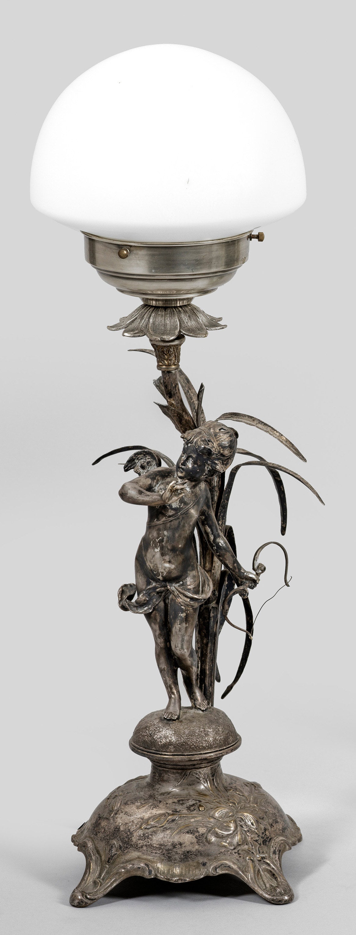 Belle Epoque-Tischlampe: Metall, versilbert und weißes Glas. Über geschweift ansteigendem, floral reliefiertem Sockel vollplastisch gestaltete Amorette sowie schlanker Schaft mit Schilfblatt als Träger für Leuchtmittel mi