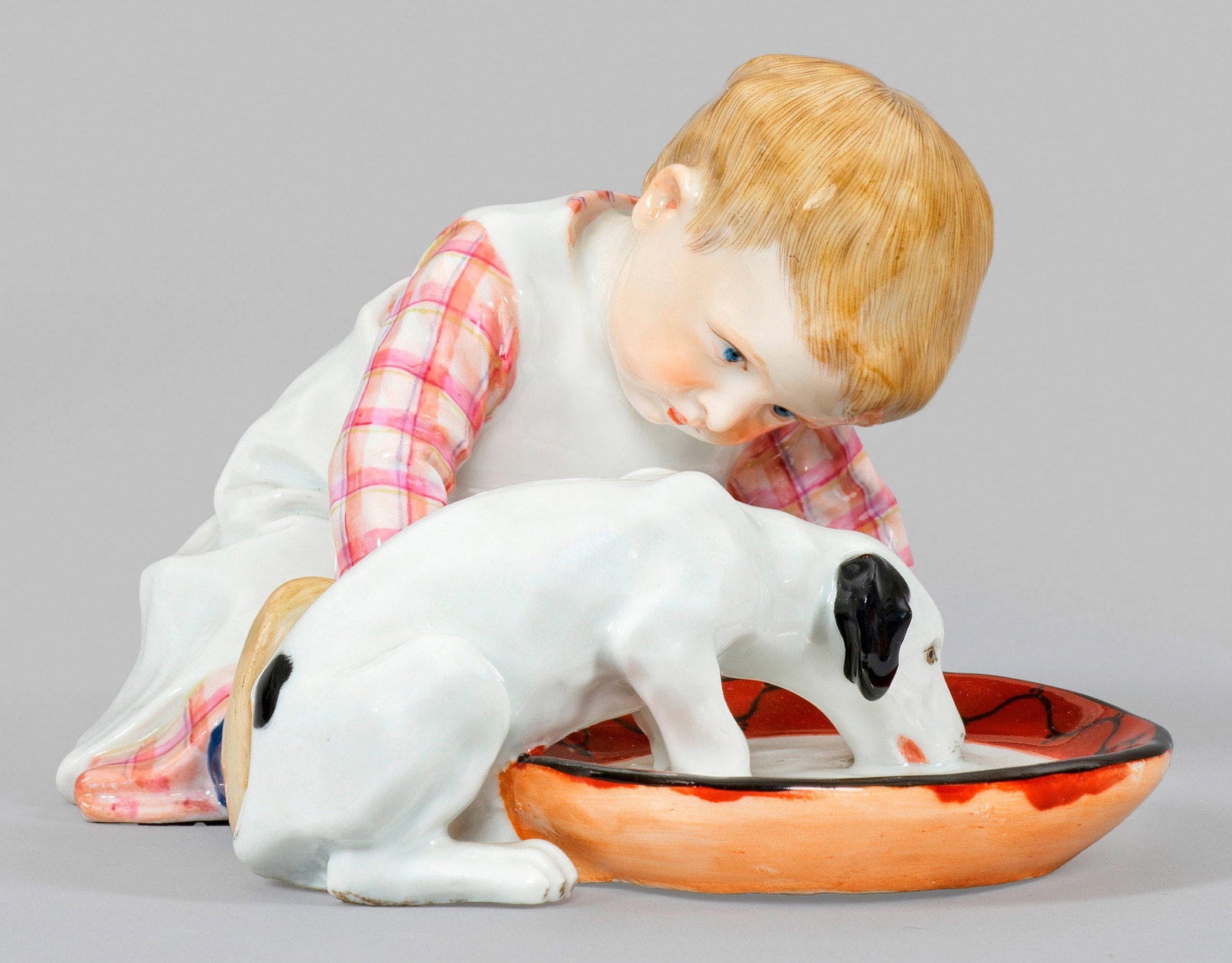 Kind mit Hund: aus der Serie der sog. Hentschelkinder. Auf dem Boden sitzendes Kind, einen kleinen Hund beobachtend, der aus einem flachen, großen Napf Milch trinkt. Polychrome Aufglasurmalerei. Entw. Konrad Hentsc