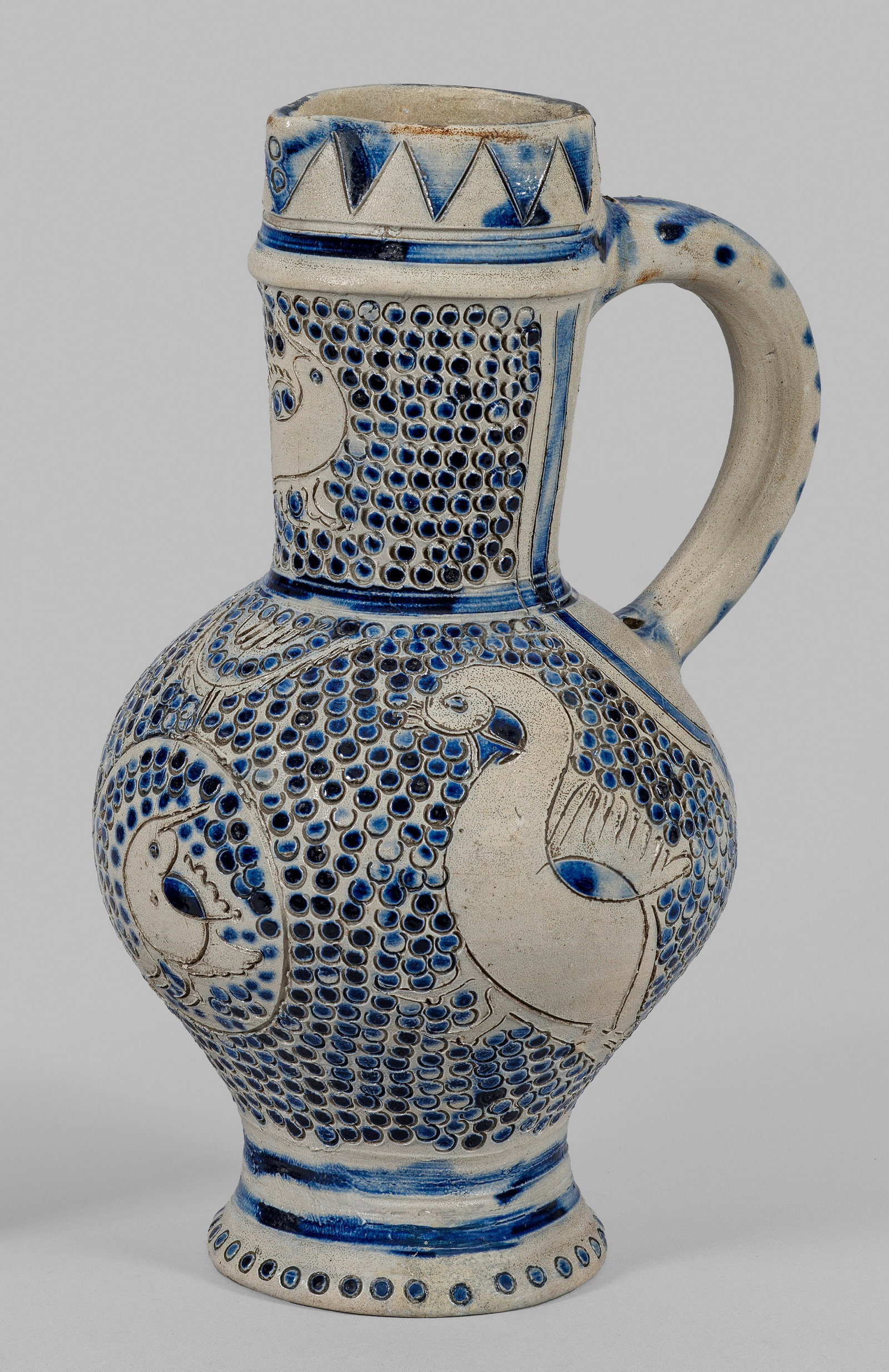 Enghalskanne: Salzglasiertes, hellgraues Steinzeug. Hoher, profilierter Fuß, ovoider Korpus, abgesetzter, zylindrischer Hals mit kurzer Schnaupe. Ritzdekor aus Vögeln in Redtechnik, der Fond mit gestempeltem Schu