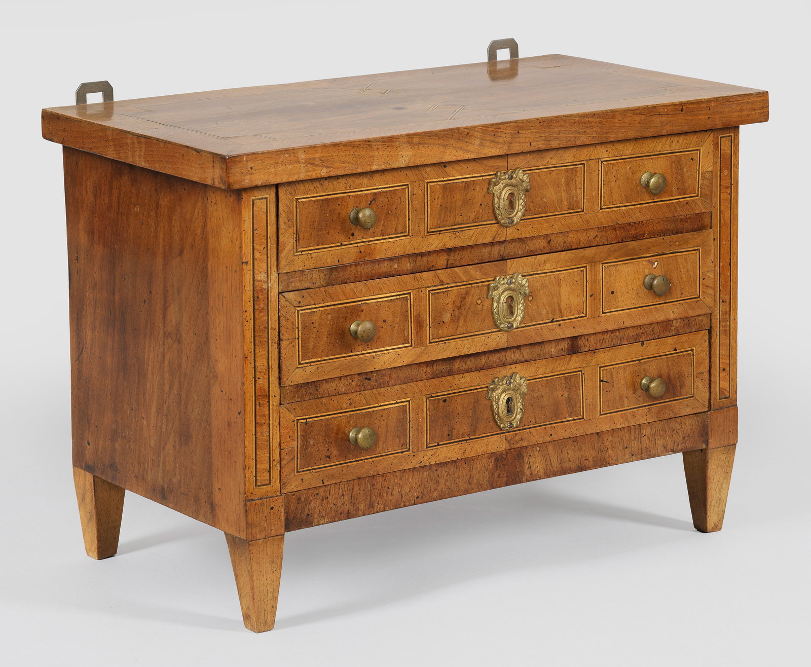 Louis XVI-Modellkommode: Nussbaum, furniert sowie schmale Fadeneinlagen. Gerader, querrechteckig angelegter Korpus auf konischen Vierkantfüßen. In der gegliederten Front drei Schubladen. Gering überstehende Deckplatte. H.