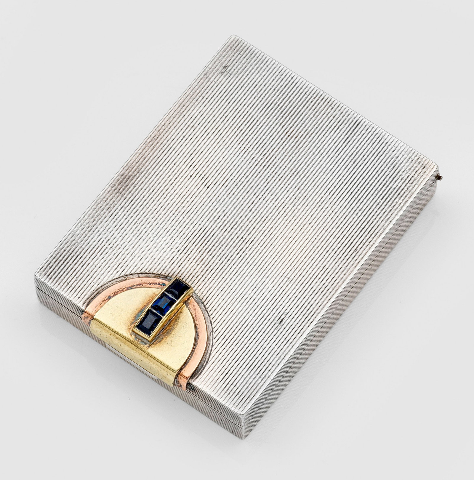 Art Déco-Puderdose von Cartier: Sterlingsilber und Gold, 14 ct.; Rechteckige Grundform mit flächendeckendem Dekor aus feinen Rillen, Schauseitig mit drei blauen Steinen besetzter halbkreisförmiger Drücker aus Gelbgold mit abgestu