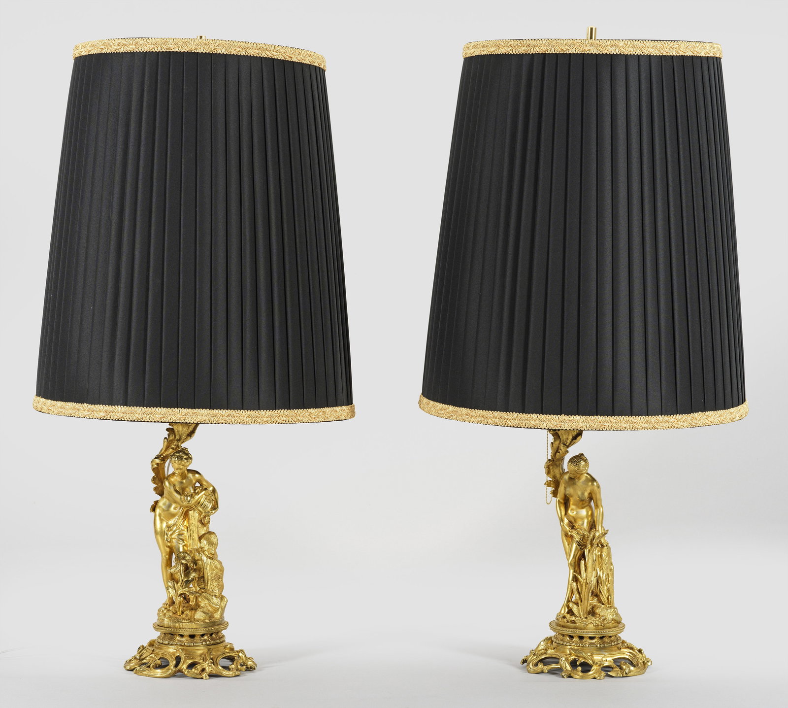 Paar Belle Epoque-Tischlampen: 2-flg.; Bronze, vergoldet. Über durchbrochen gearbeiteter Basis aus üppigen Blattranken vollplastische Darstellung von unterschiedlichen Nymphen. Dahinter gestreckte Blattranke sowie schlanker Schaf