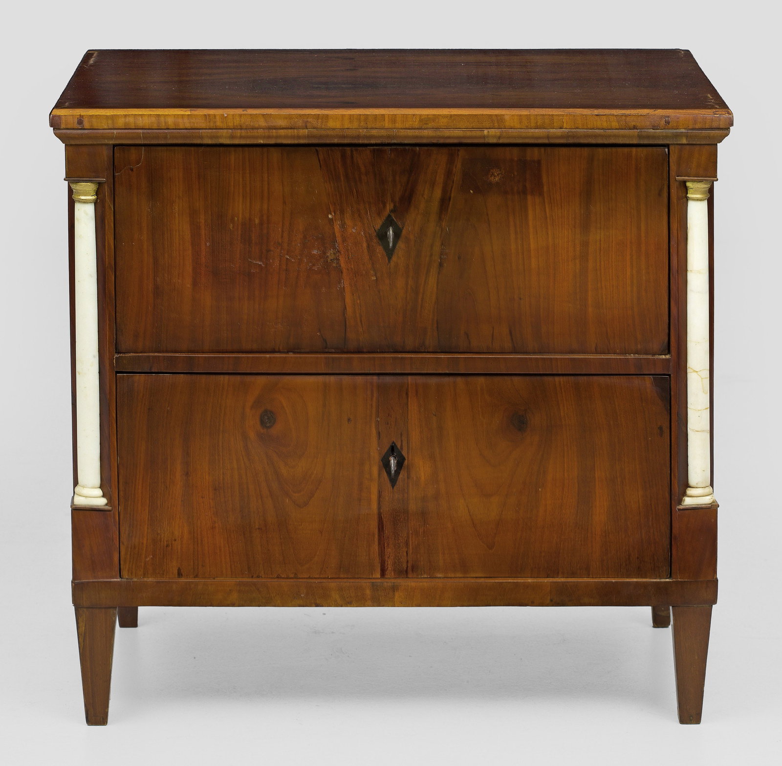 Biedermeier-Pfeilerkommode: Mahagoni, furniert sowie Alabaster, teilw. vergoldet. Gerader, rechteckig angelegter Korpus, flankiert von eingestellten Halbsäulen auf konischen Vierkantbeinen. In der Front zwei große Schubladen.