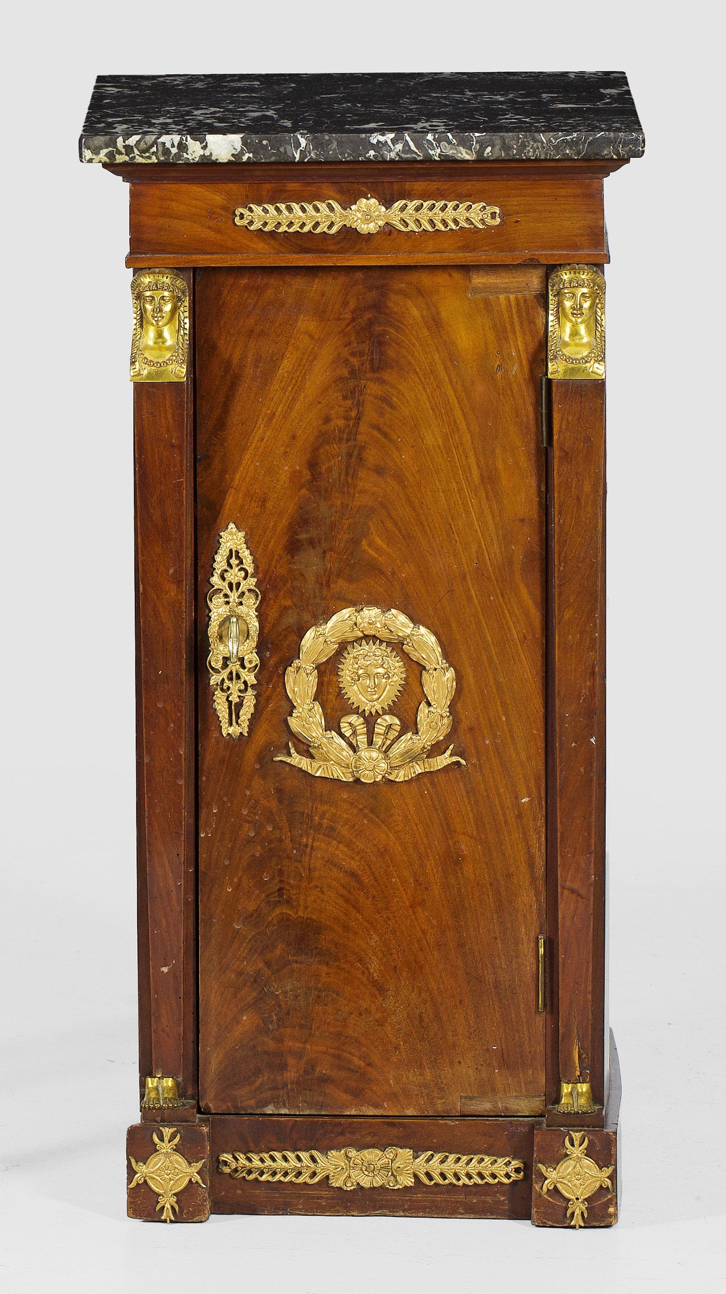 Empire-Pfeilerschrank: Mahagoni, furniert. Gerader, hochrechteckiger Korpus auf gekantetem Sockel. In der gegliederten Front schmale Tür, flankiert von eingestellten ägyptisierenden Karyatiden. Deckplatte aus grau-weiß