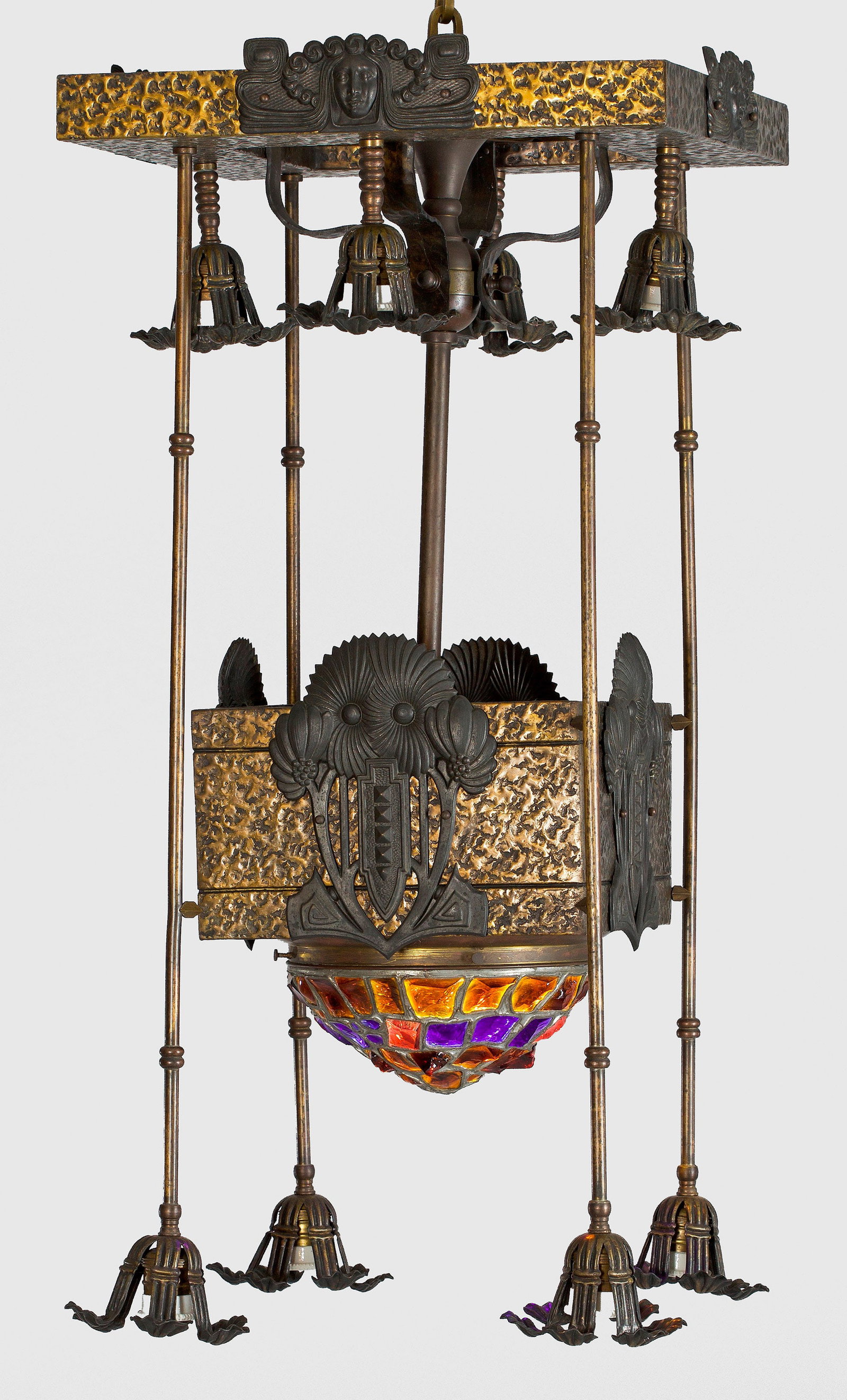 Jugendstil-Deckenlampe: 9-flg.; Messing, patiniert und farbige Glasbrocken. Quadratisches Plafon mit erhaben reliefierten Maskarons. Unterhalb davon ausgehend vier blütenförmige Lampenfassungen sowie schmale Rundstäbe fü