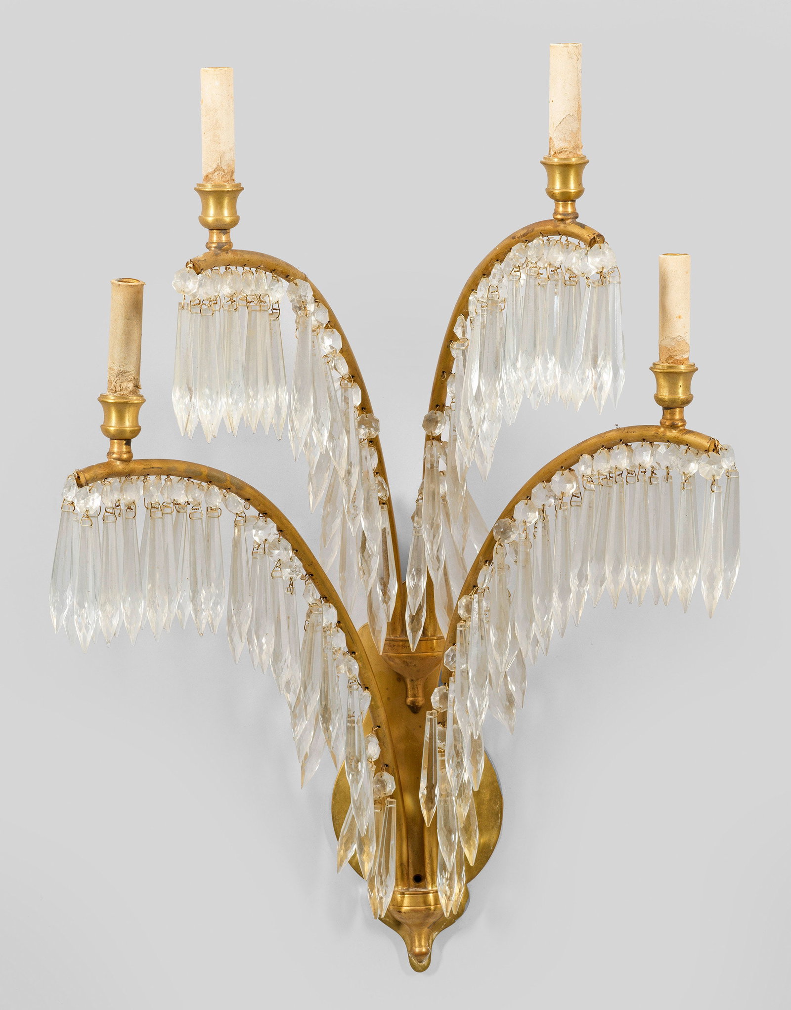 Wandleuchter: 4-flg.; Messing und farbloses Glas. Flaches, balusterförmiges Wandschild. Davon ausgehend vier stark gebogte Leuchterarme mit dicht angeordnetem Prismenbehang als Träger für Leuchtmittel. Elektrifi