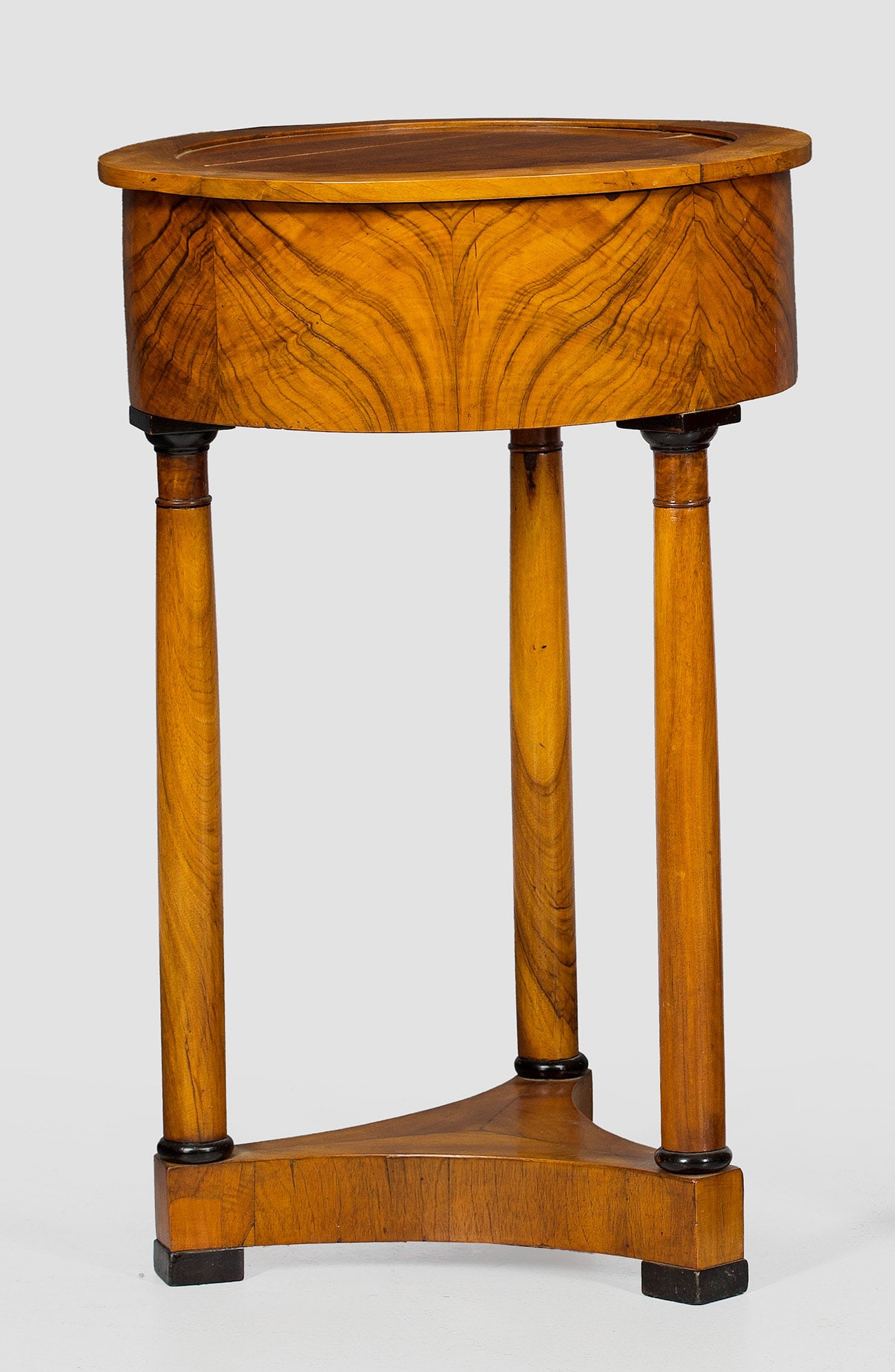 Biedermeier-Trommeltischchen: Nussbaum, furniert. Über dreiseitig konkav eingezogener Sockelplatte konische Vollsäulen als Träger für bombierten Zargenkasten sowie runde, aufklappbare Deckplatte. H. 75 cm. D. 50 cm.