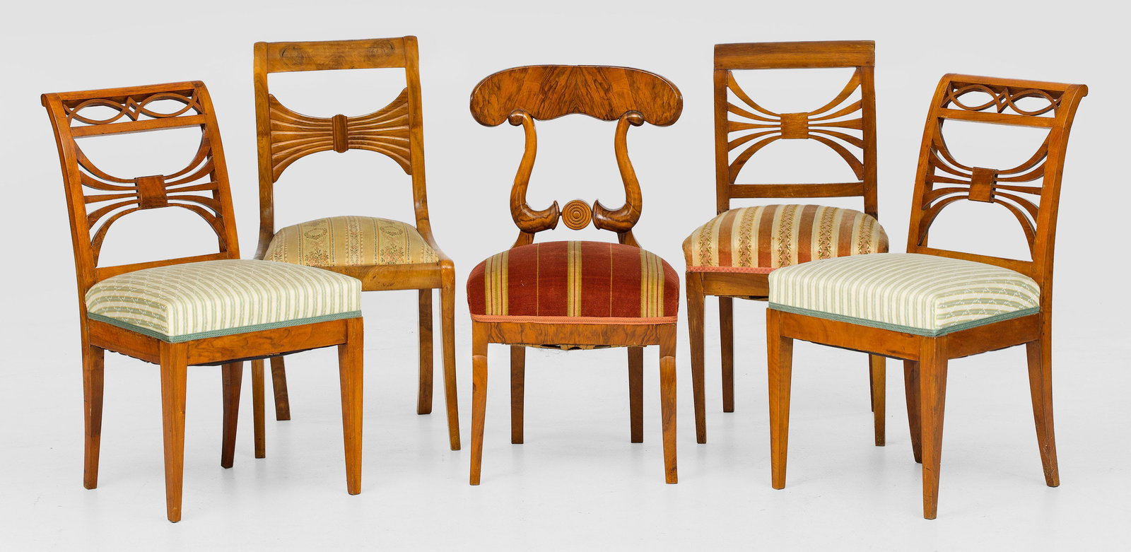 Biedermeier-Stuhlgruppe: 5-tlg.; Nussbaum, massiv und furniert. Trapezförmige, teilw. bombierte Zarge auf konischen, teilw. geschwungenen Beinen. Unterschiedliche Rückenlehnengestaltungen. Sitzfläche gepolstert und bezogen