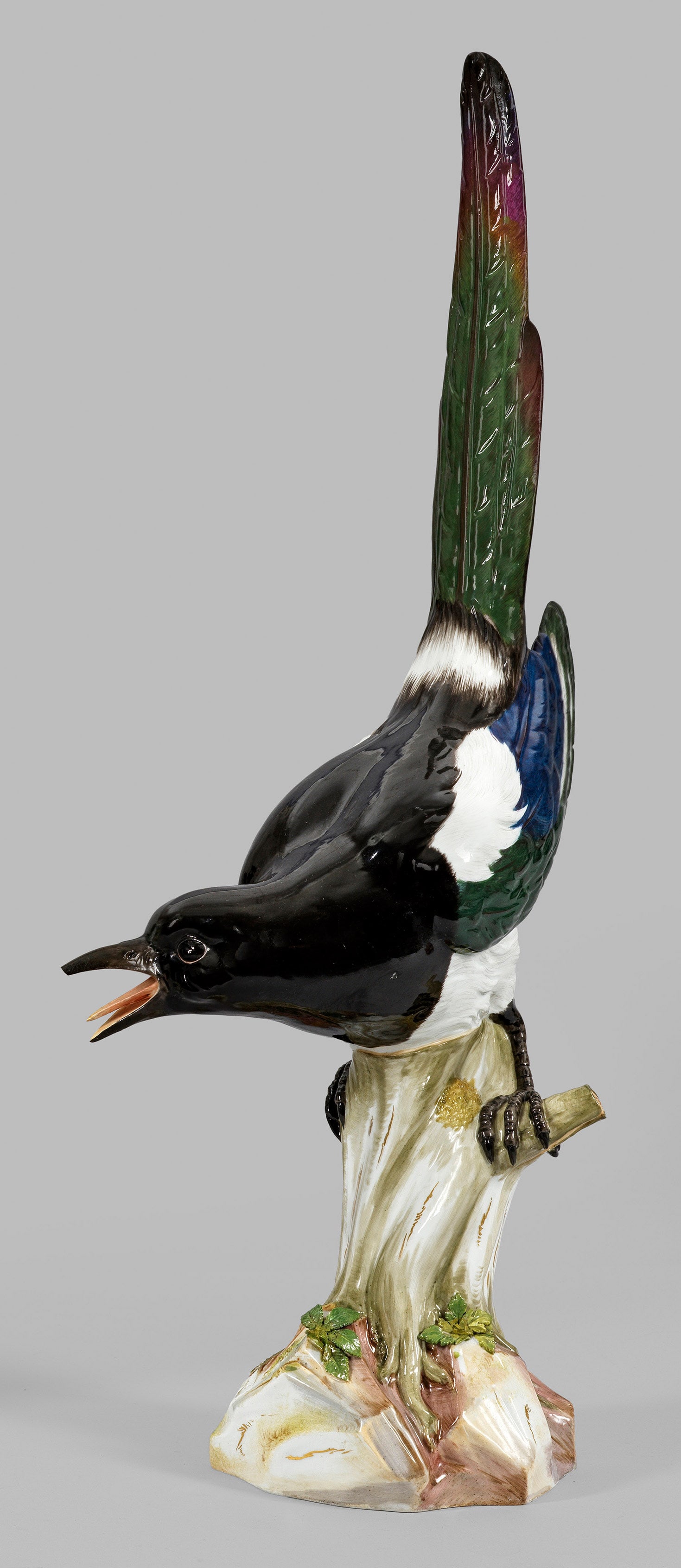 Große Elster: Auf einem mit Blättern belegten Baumstamm sitzende Elster mit naturalistisch gestaltetem Gefieder und weit geöffnetem Schnabel. Polychrome Malerei mit dezenter Goldstaffage. Entw. Johann Joachim Kae