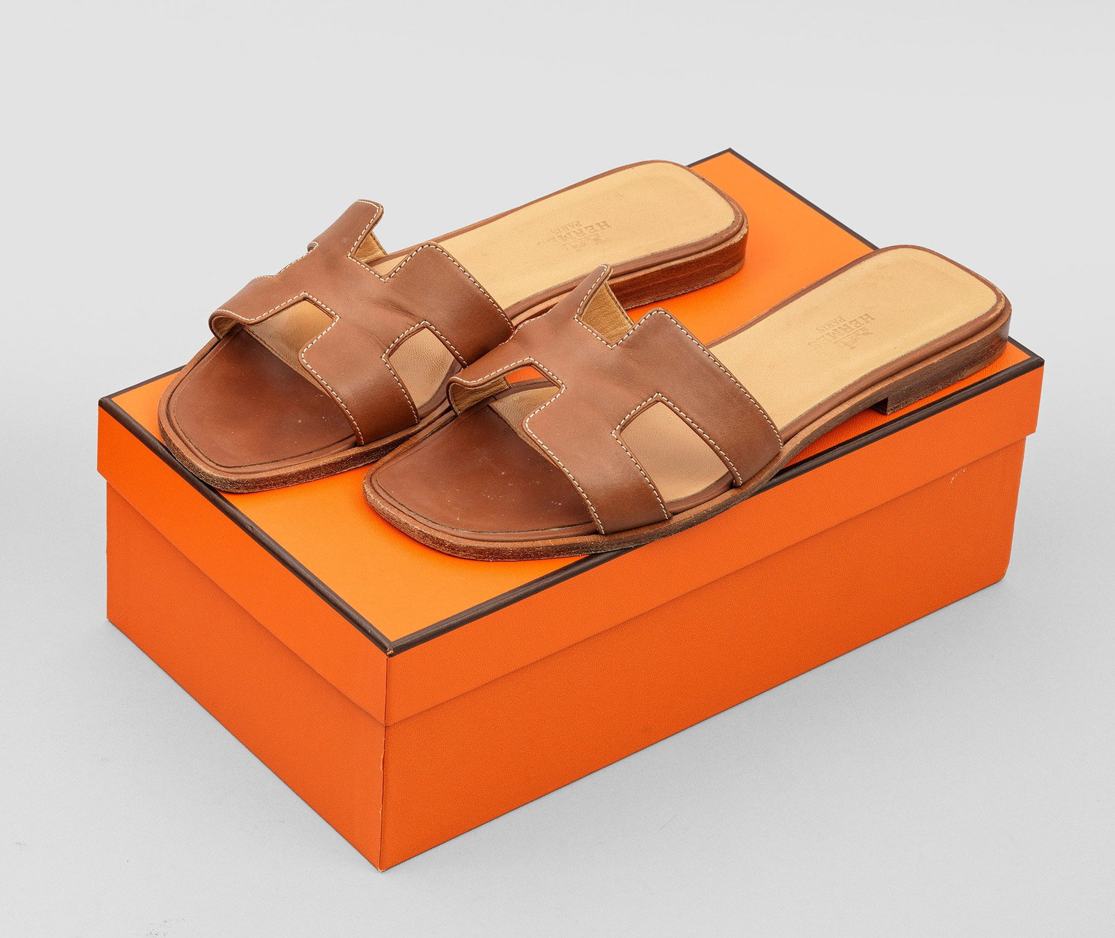 Paar Damen-Sandalen "Oran" von HERMÈS: Cognacfarbenes Leder. Flache Sandale mit breitem Riegel aus stilisiertem H-Logo. Gr. 38. Getragen. Original Box und Staubbeutel. A pair of pre-owned brown leather sandals by HERMÈS. Worn. Box and dus