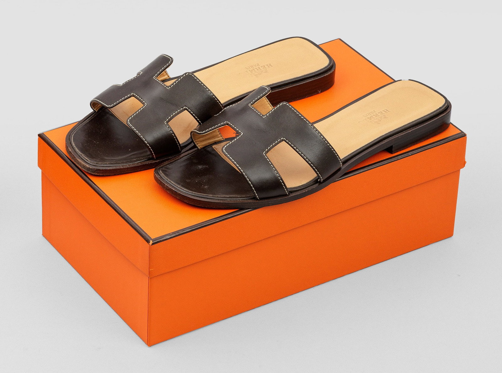 Paar Damen-Sandalen "Oran" von HERMÈS: Dunkelbraunes, ébènefarbenes Leder. Flache Sandale mit breitem Riegel aus stilisiertem H-Logo. Gr. 38. Getragen. Original Box und Staubbeutel. A pair of pre-owned dark brown leather sandals by HERM�