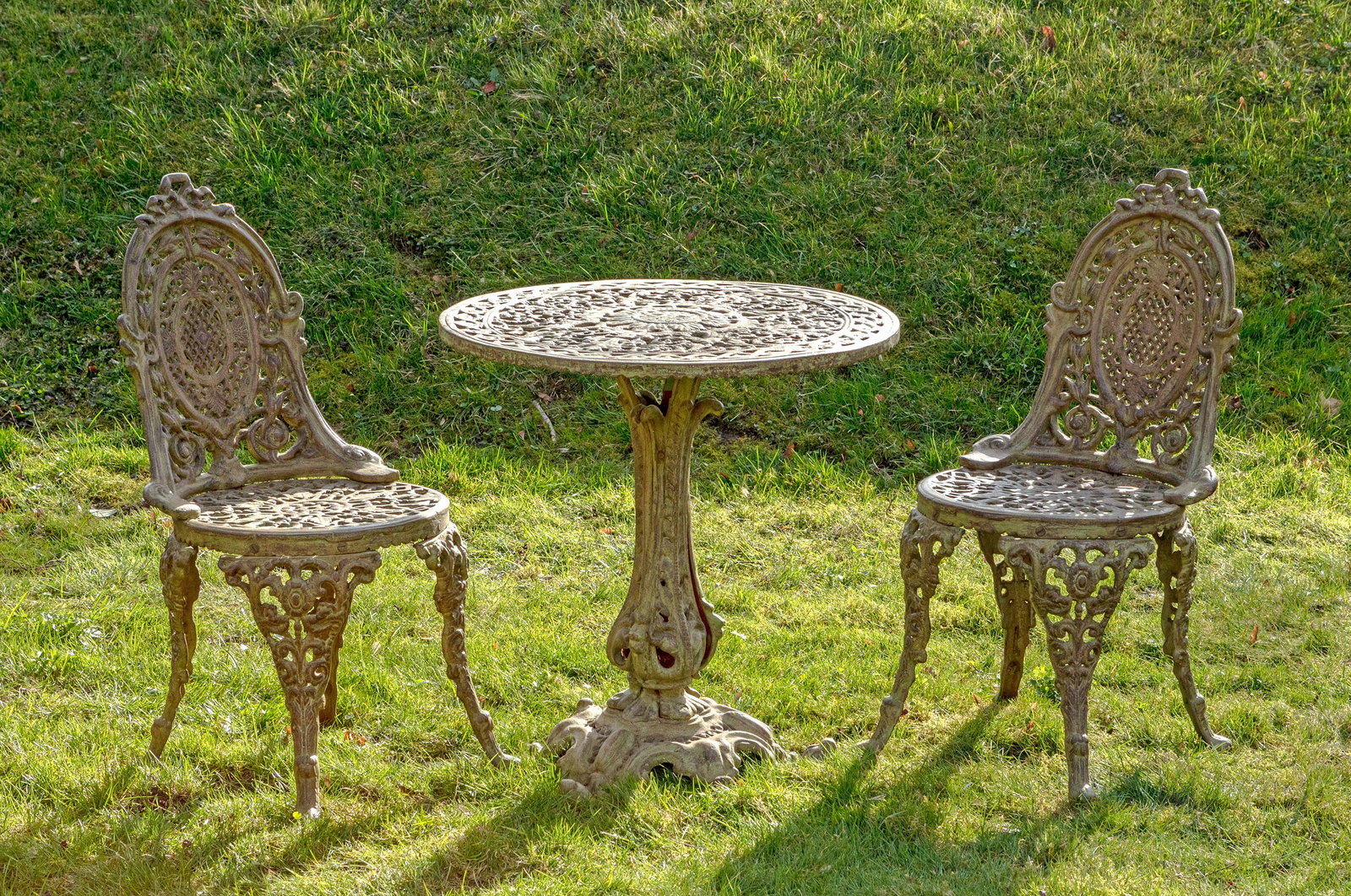 Garnitur Gartenmöbel im Belle Epoque-Stil: 3-tlg.; Tisch und Paar Stühle. Gusseisen, teilw. hellgrün lackiert. Balusterförmiger Schaft auf breiten Voluten als Träger für runde, durchbrochen gearbeitete Deckplatte. Korrespondierende Stühl