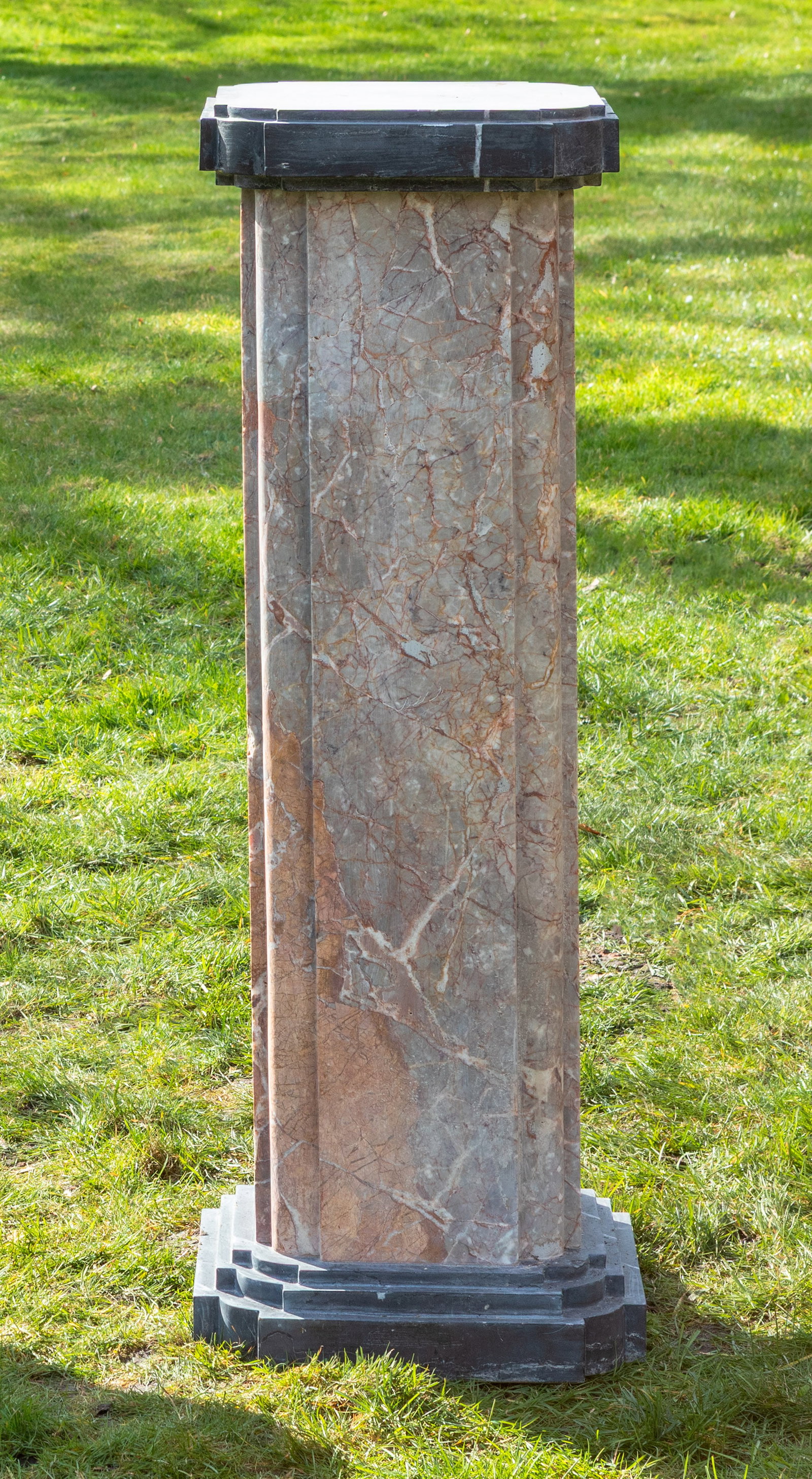 Podestsockel: In unterschiedlichen Cremetönen gemaserter sowie dunkelgrauer Marmor. Über gestuft ansteigender Basis, kräftiger, gekanteter Schaft mit gerundeten Ecken als Träger für korrespondierende, profilie