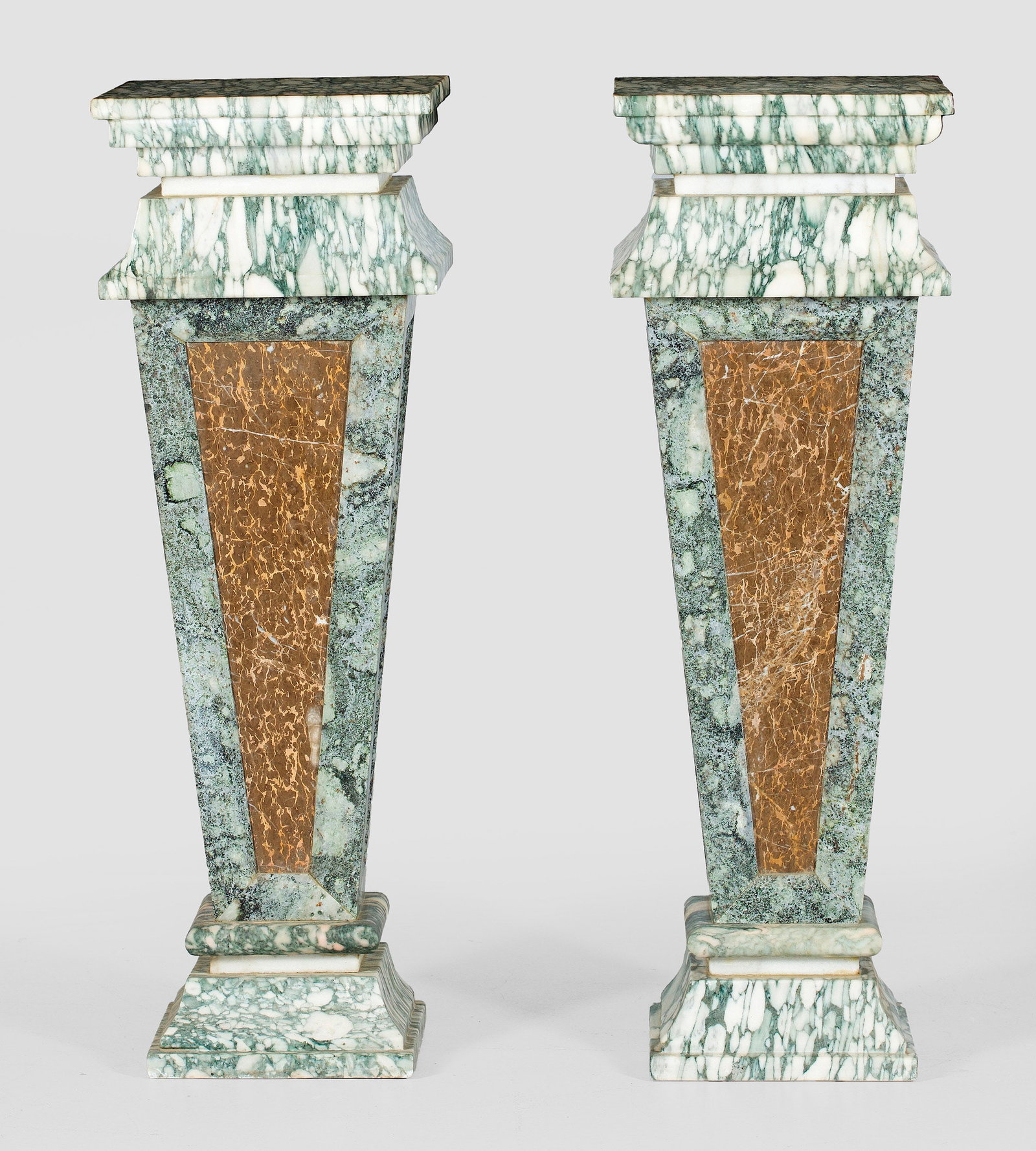 Paar Skulpturensäulen: Weißer, grau-grün sowie in unterschiedlichen Beigetönen gemaserter Marmor. Über geschweift ansteigender Basis kräftiger, sich konisch weitender Schaft mit flach zurückgesetzten Füllungen. Korre