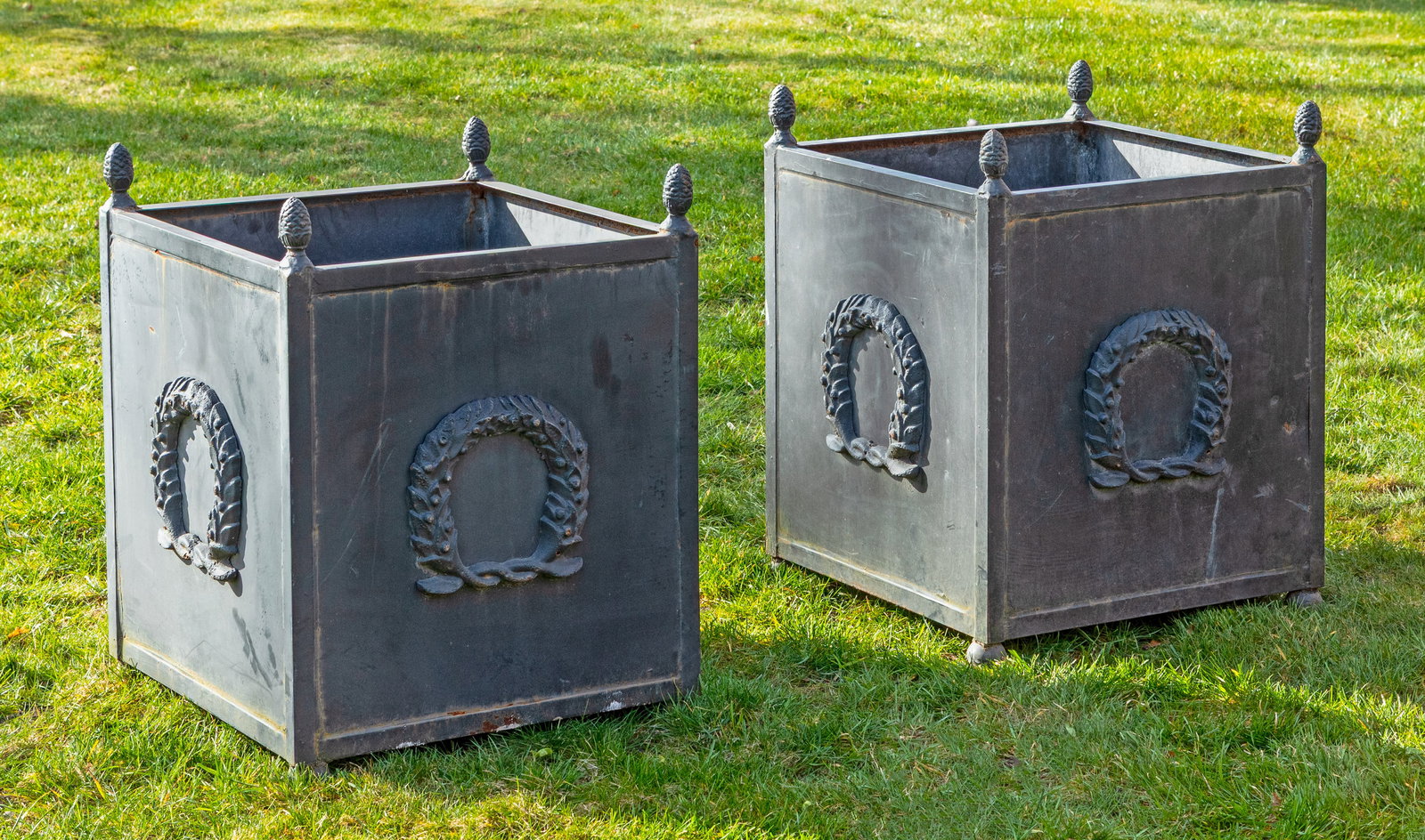 Zwei Pflanzgefäße: Metall, anthrazitfarben lackiert. Gerader, quadratisch angelegter Korpus mit erhaben reliefierten Kranzmotiven sowie Bekrönungen in Form von Pinienzapfen. H. 70 cm. 54 cm x 54 cm. A pair of French gr