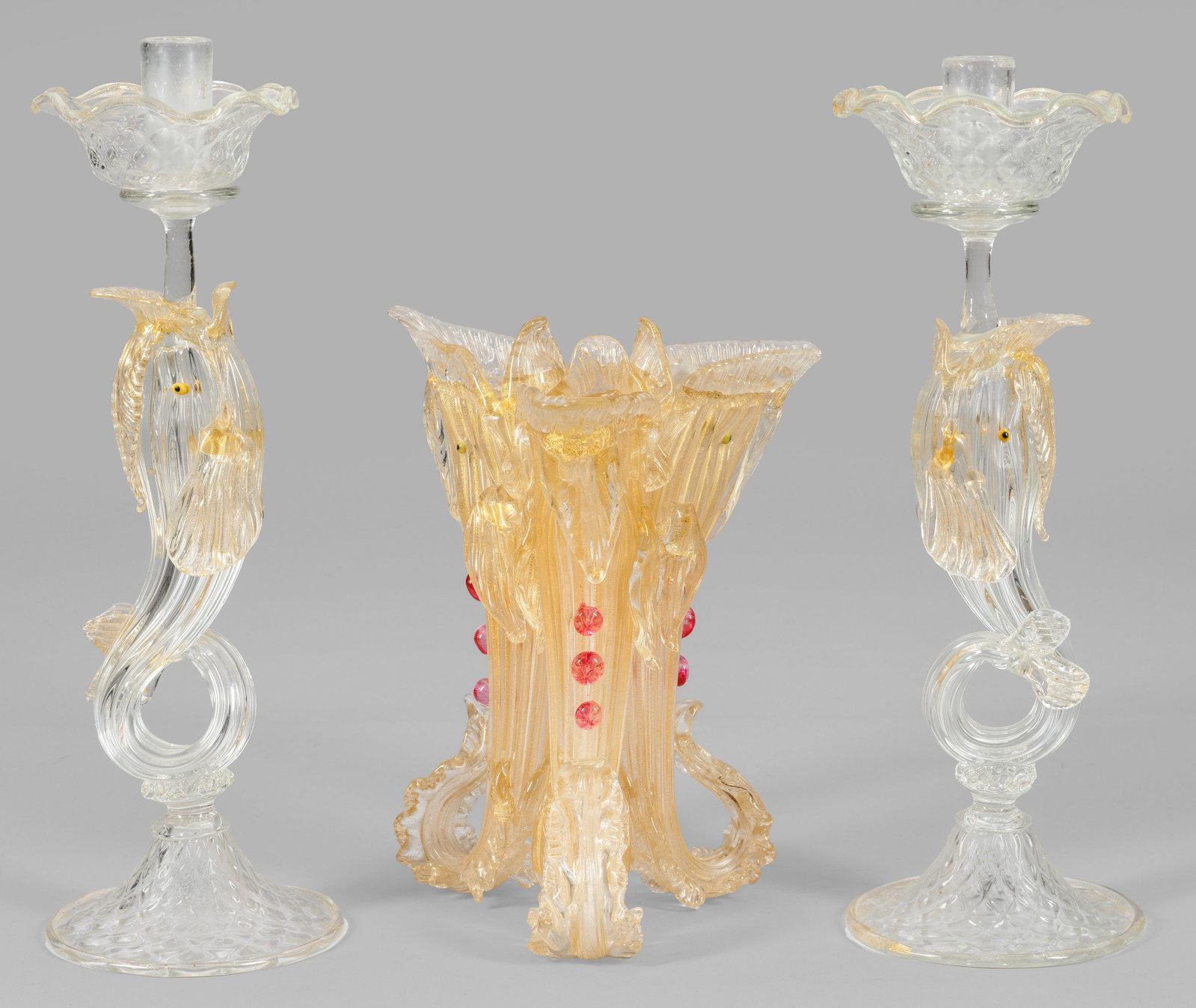 Prachtvolle Murano-Tafeldekoration mit Delfin-Dekor: 3-tlg.; Farbloses und rosafarbenes Glas, teilw. mit Einschmelzungen aus fein zersprengter Goldfolie, die Augen aus gelbem und schwarzem Glas. Zwei 1-flg. Zierkerzenleuchter mit plastischen Delfinschä