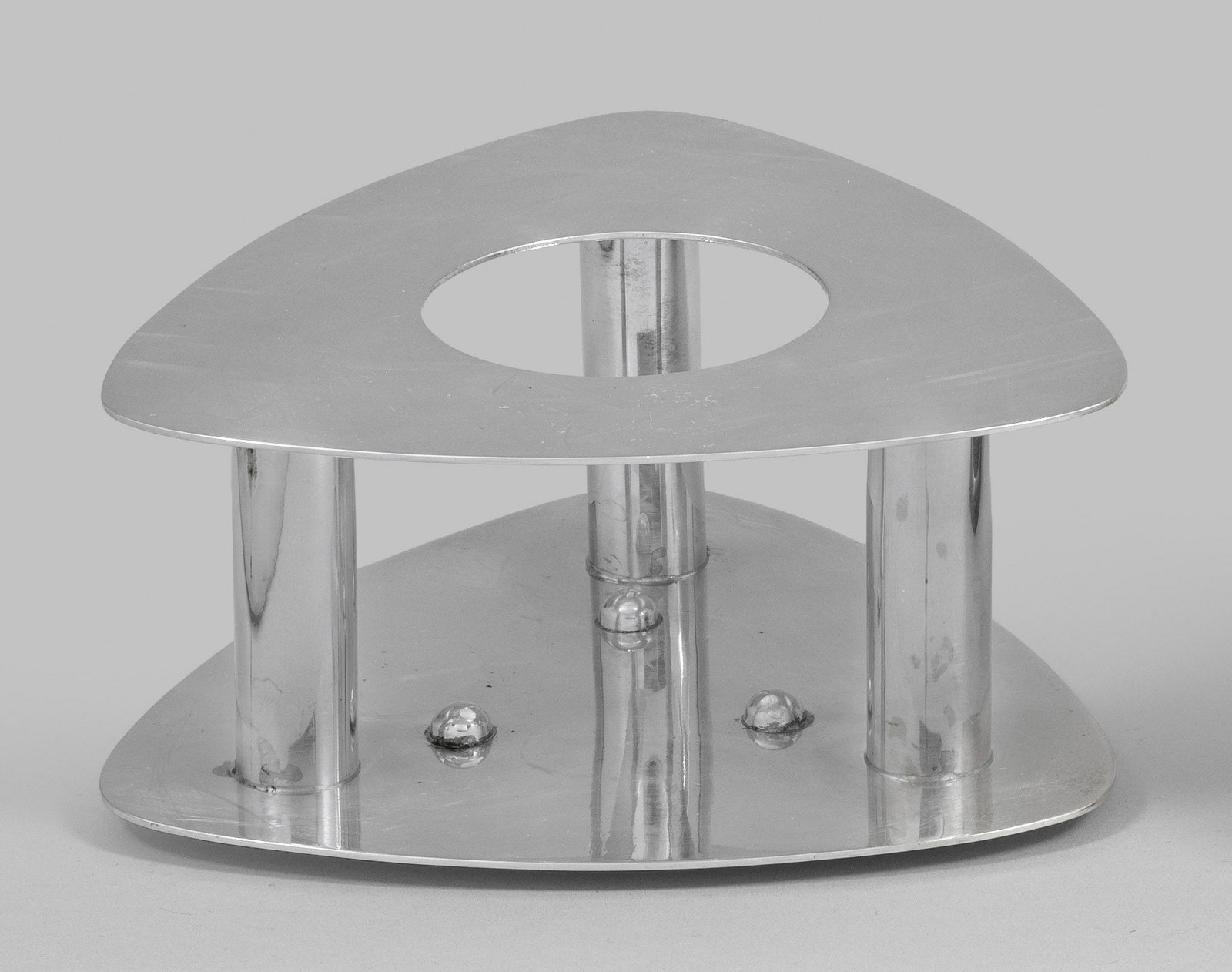 Stövchen: Silber. Auf dreieckiger Unterplatte mit Standnoppen und gerundeten Ecken drei zylindrische Säulen. Darauf korrespondierende dreieckige Platte mit mittiger, runder Aussparung. Gest. "Handmade in silve