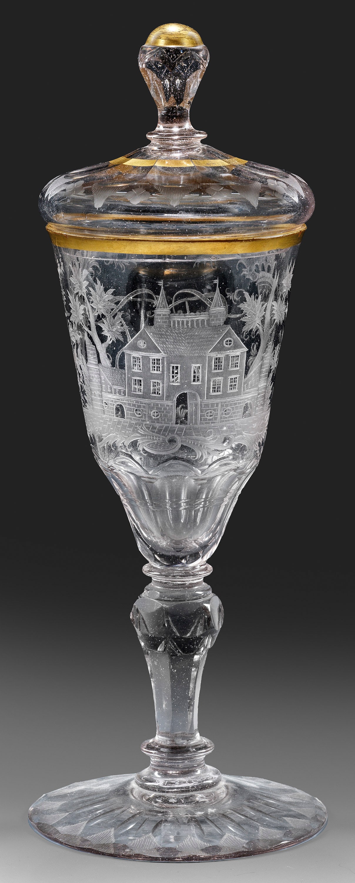 Schlesischer Deckelpokal mit Monogramm und Schlossdekor: Farbloses, leicht graustichiges Glas, geschliffen, goldgerändert. Flacher Scheibenfuß mit Bodenrosette, auf der Oberseite eine Bordüre aus mandelförmigen Schliffbögen und schraffierten Dreiecken.
