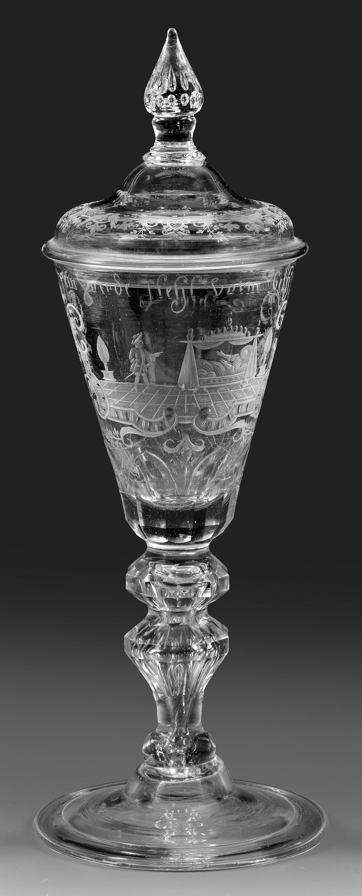 Großer Deckelpokal mit erotischer Szene und Sinnspruch: Farbloses Glas mit Abriss, partiell geschliffen. Konische Kuppa, geschälter Ansatz, facettierter Balusterschaft, mittig ansteigender Fuß mit umgeschlagenem Rand. Matt geschnittener, teilw. geblänkt