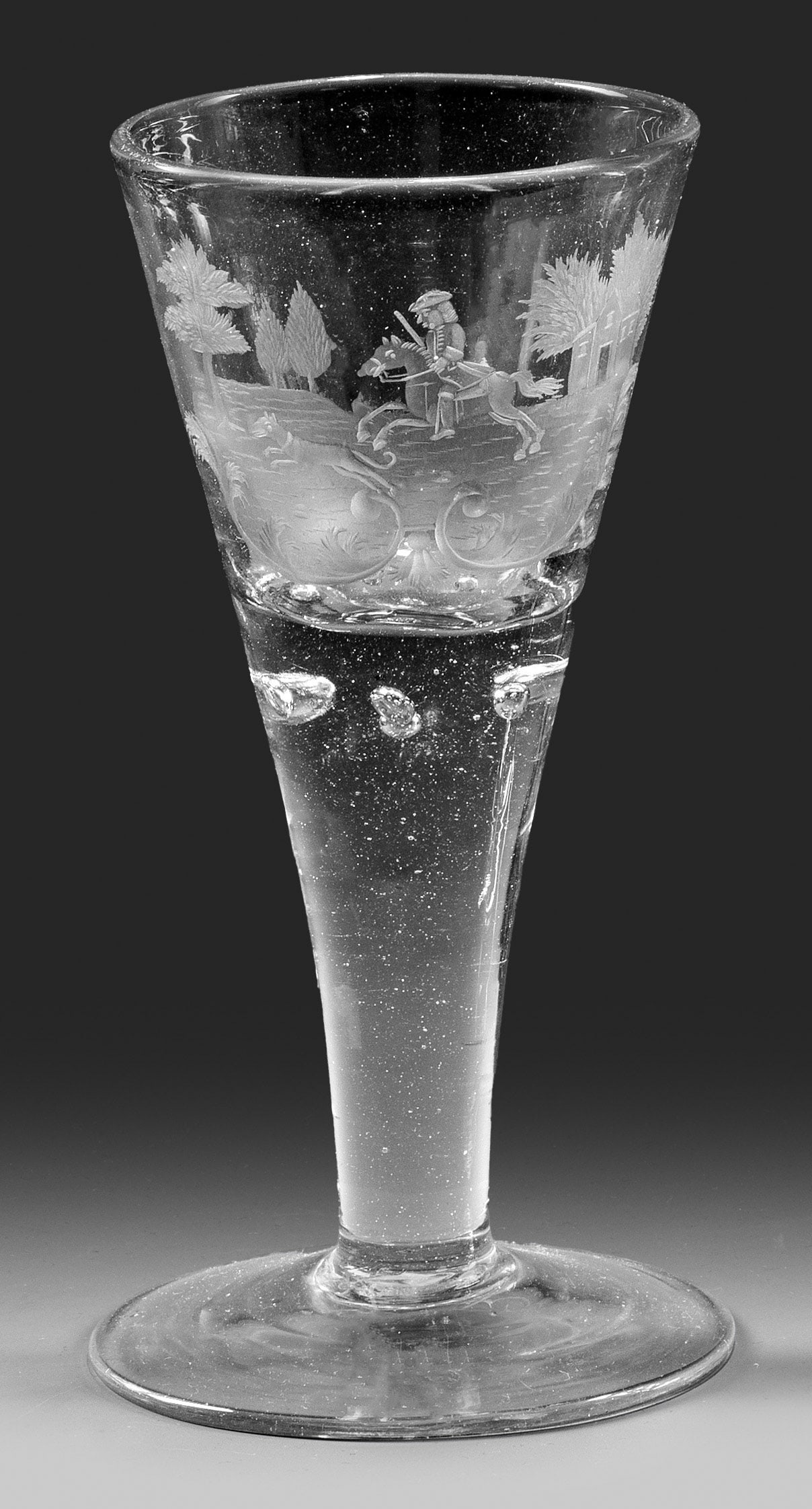 Spitzkelch-Pokal mit Jäger zu Pferde: Farbloses Glas, im Schaft 6 kleine eingestochene Luftblasen. Auf der Kuppa geschnittener Dekor eines berittenen Jägers mit seinem Hund in stilisierter Landschaft. H. 16,8 cm. A North German or Hessia