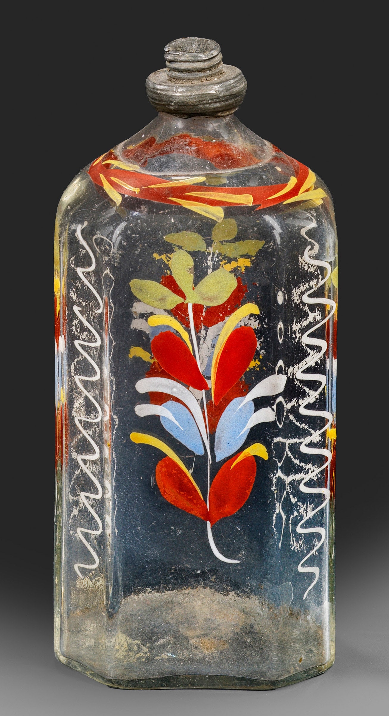 Schnapsflasche (Branntweinflasche): Farbloses, leicht graustichiges Glas mit Abriss im Boden. Querrechteckige Form mit abgeschrägten Ecken, gerundeter Schulter und zinnmontierter Mündung. Polychromer Dekor in opaker Emailmalerei aus f