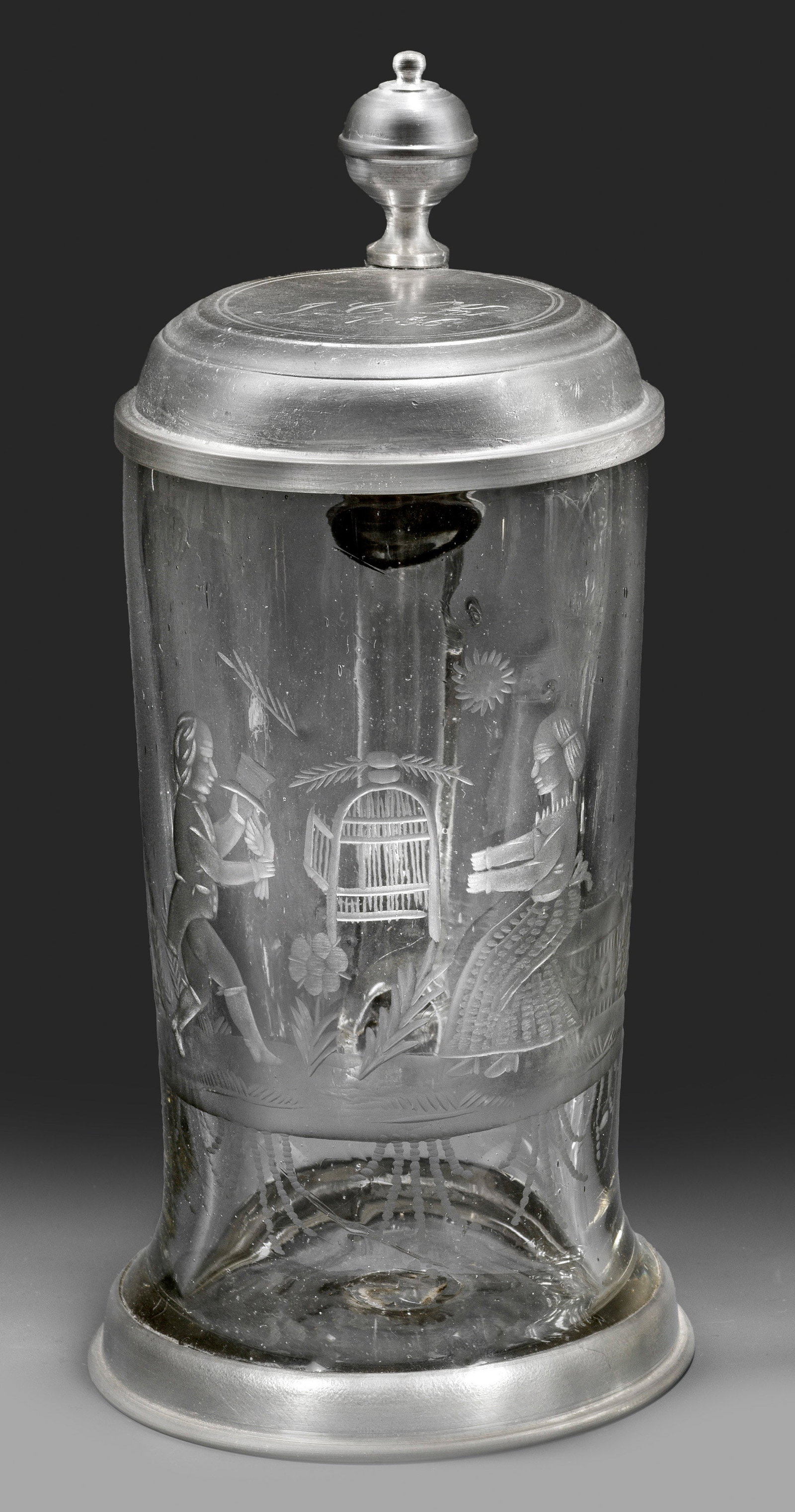 Biedermeier-Hochzeitskrug mit jungem Liebespaar: Farbloses, leicht graustichiges Glas, im Boden Abriss. Hohe, schlanke Walzenkrugform, der Bandhenkel mit volutiertem Ansatz. Schauseitig geschnittener szenischer Dekor: auf einem Landschaftssockel sit