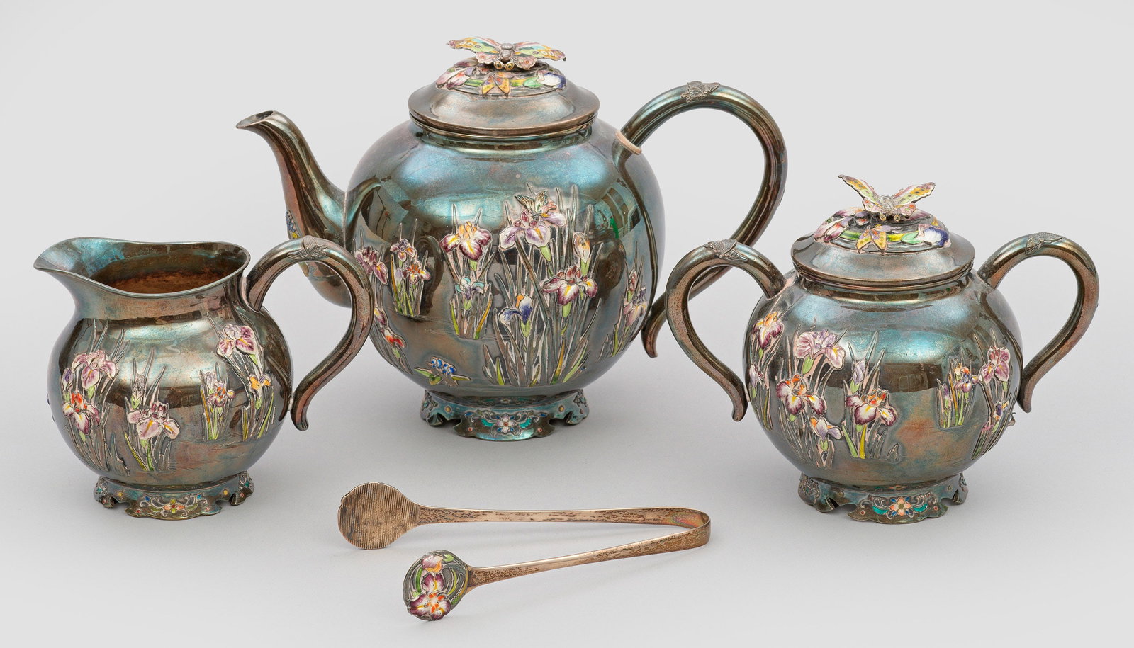 Japanisches Teeservice mit Schwertlilien-Dekor: Silber, teilw. emailliert. 4-tlg.; Teekanne, Sahnegießer, Zuckerdose, Zuckerzange. Auf rundem Fuß kugeliger Korpus mit beidseitigem, polychrom emaillierten Reliefdekor aus Schwertlinien und anderen 