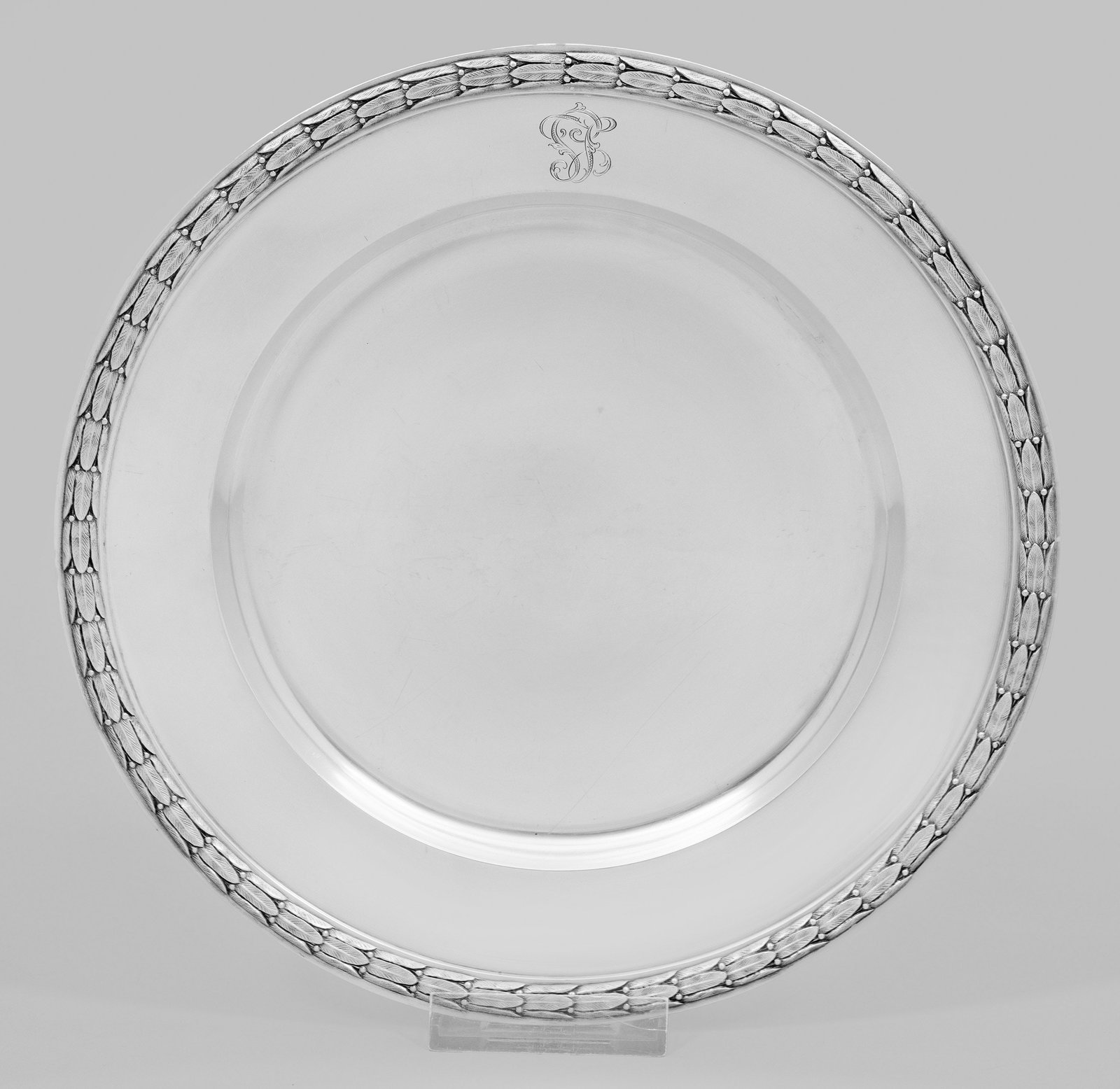 Platzteller mit Lorbeerdekor: Silber. Runde Grundform mit leicht vertieftem Spiegel und leicht ansteigender, am Rand von Lorbeerrelief gesäumter Fahne mit ligiertem Monogramm. Gest. 84, Stadtmarke, Herstellerzeichen. Gew. ca. 590