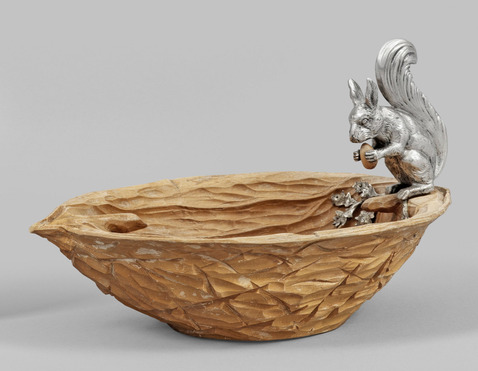Eichhörnchen auf Nussschale: Silber und Holz. Plastisch ausgearbeitete Holzschale in Form einer Walnusshälfte. Auf dem Rand sitzendes, silbernes Eichhörnchen mit einer Haselnuss in den Vorderpfoten. Daneben aufgelegt zwei Äste