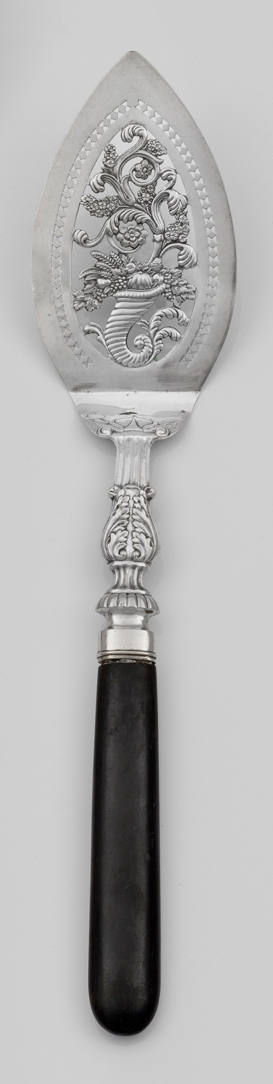 Fischheber mit Füllhorn: Silber. Spitz zulaufende, durchbrochen gearbeitete und reliefierte Laffe. Zentral die Darstellung eines Füllhorns, aus dem ein Baum wächst. Leicht konischer Griff aus ebonisiertem Holz. Minim. geris