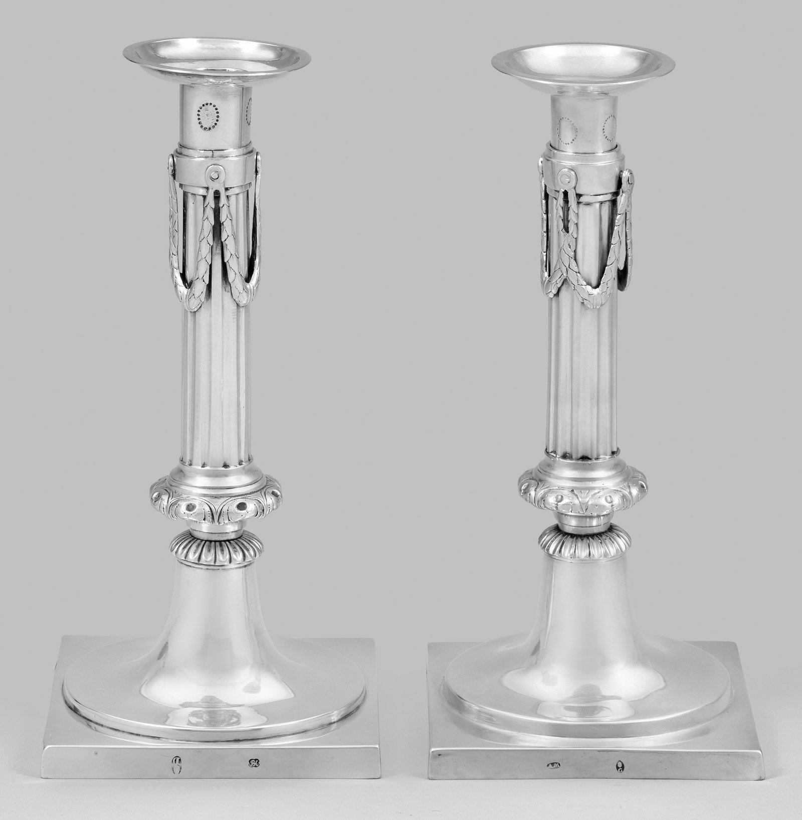 Zwei klassizistische Tafelleuchter: Silber. Quadratische Plinthe übergehend in trompetenförmigen Fuß. Aus Palmettenkranz und Volutennodus aufsteigender kannelierter Säulenschaft mit aufgelegten Lorbeerfestons. Herausnehmbare Tülle 