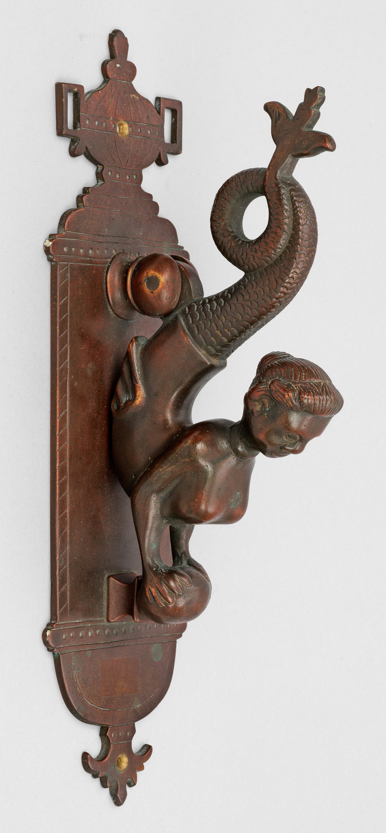 Schwerer Türklopfer: Bronze. Flaches, profiliertes Wandschild mit Vasenbekrönung. Oberhalb davon ausgehend beweglicher Türklopfer in vollplastischer Darstellung eines Meerweibchens. H. 38 cm. A North German bronze door 
