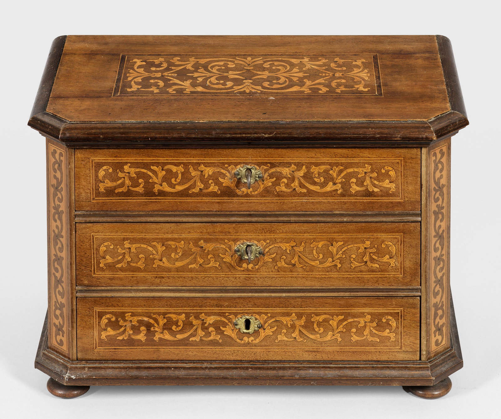 Napoleon III-Modellkommode: Nussbaum, furniert. Gerader, querrechteckig angelegter Korpus, flankiert von schräggestellten Ecklisenen auf schmaler Zarge und gedrückten Füßen. In der gegliederten Front drei Schubladen. Gering 