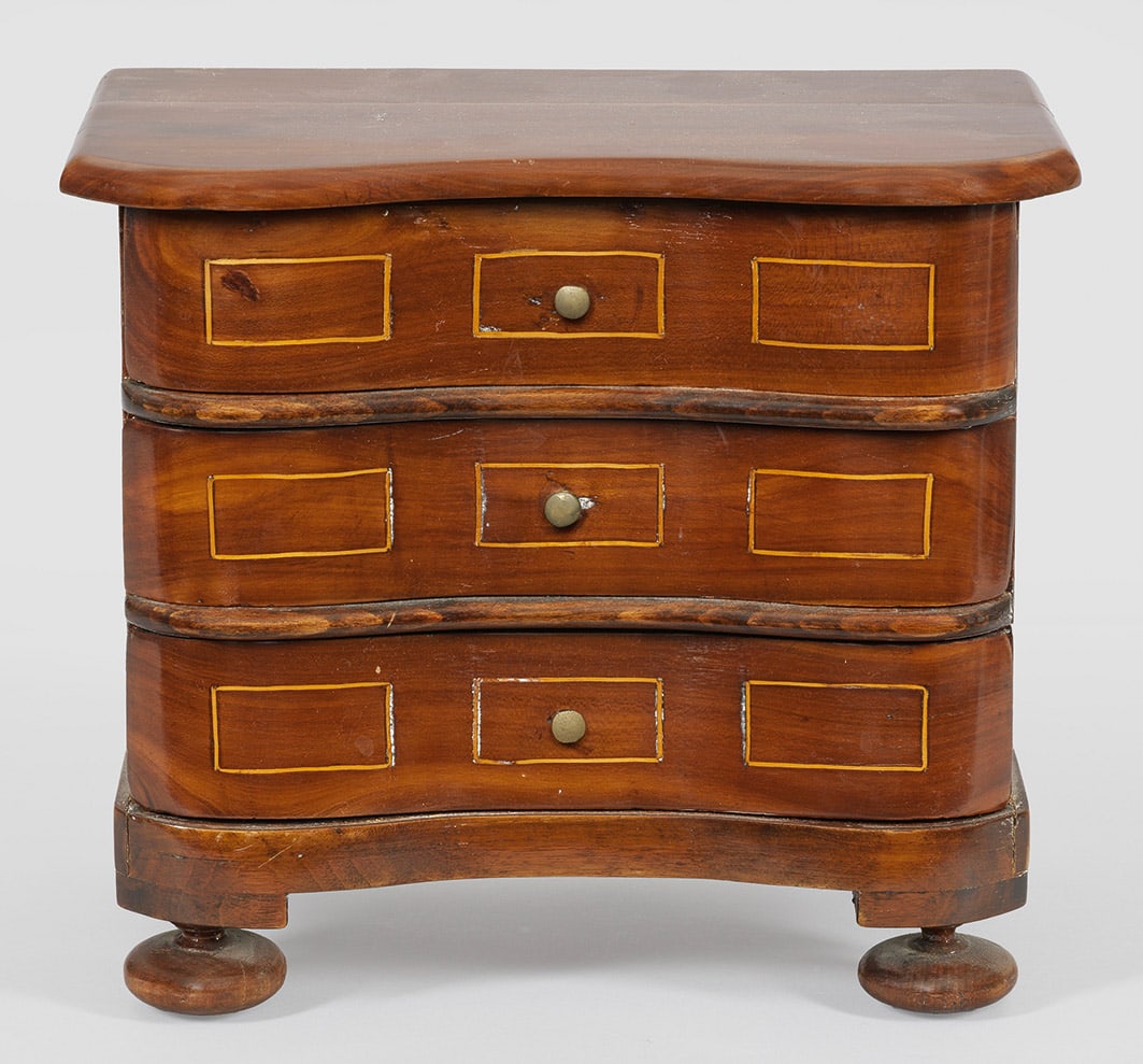Kleine Barock-Modellkommode: Nussbaum, furniert. Rechteckiger Korpus auf gedrückten Füßen. In der gegliederten Front drei geschweifte Schubladen. H. 23 cm. 27 cm x 17 cm. Provenienz: Auflösung der Privatsammlung Horst Schach,