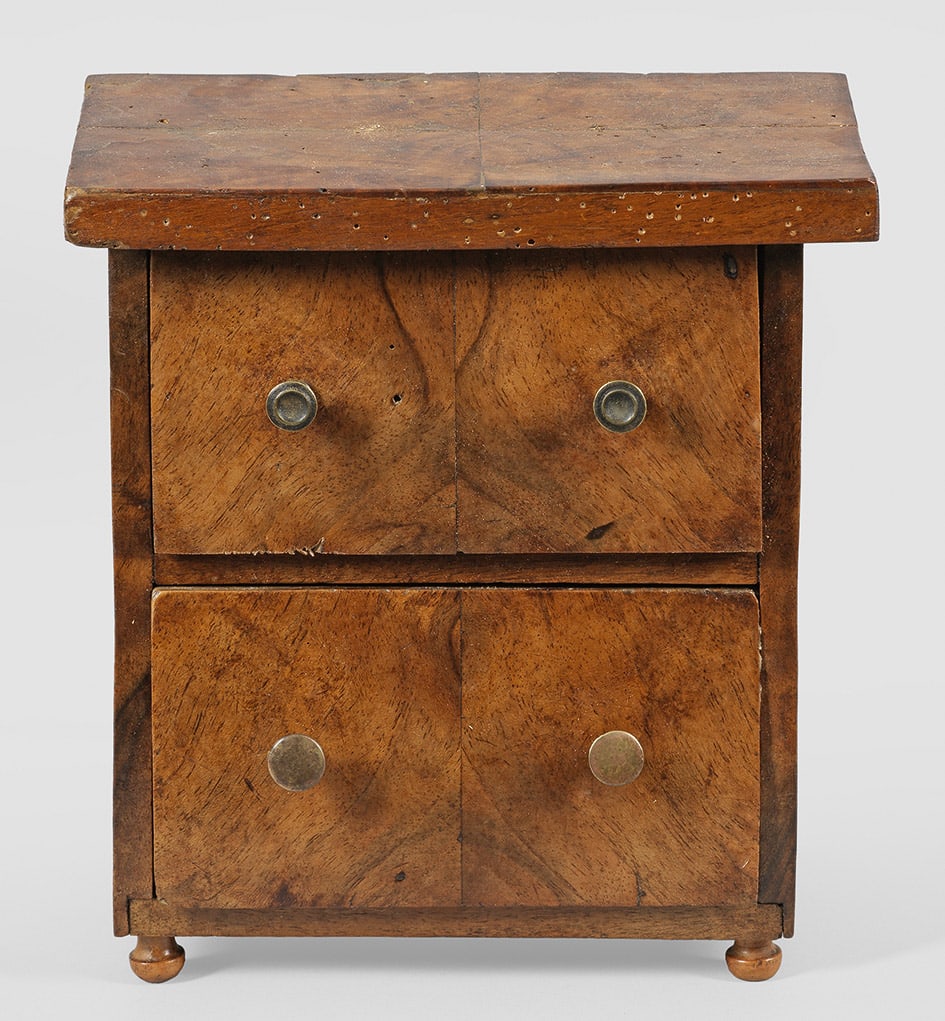 Kleine Biedermeier-Modellkommode: Nussbaum, furniert. Gerader, hochrechteckiger, zweischübiger Korpus auf kleinen Blausterfüßen. Rechteckige Deckplatte. Altersspuren und Erg.; H. 21 cm. 19,5 cm x 11 cm. Provenienz: Auflösung der P