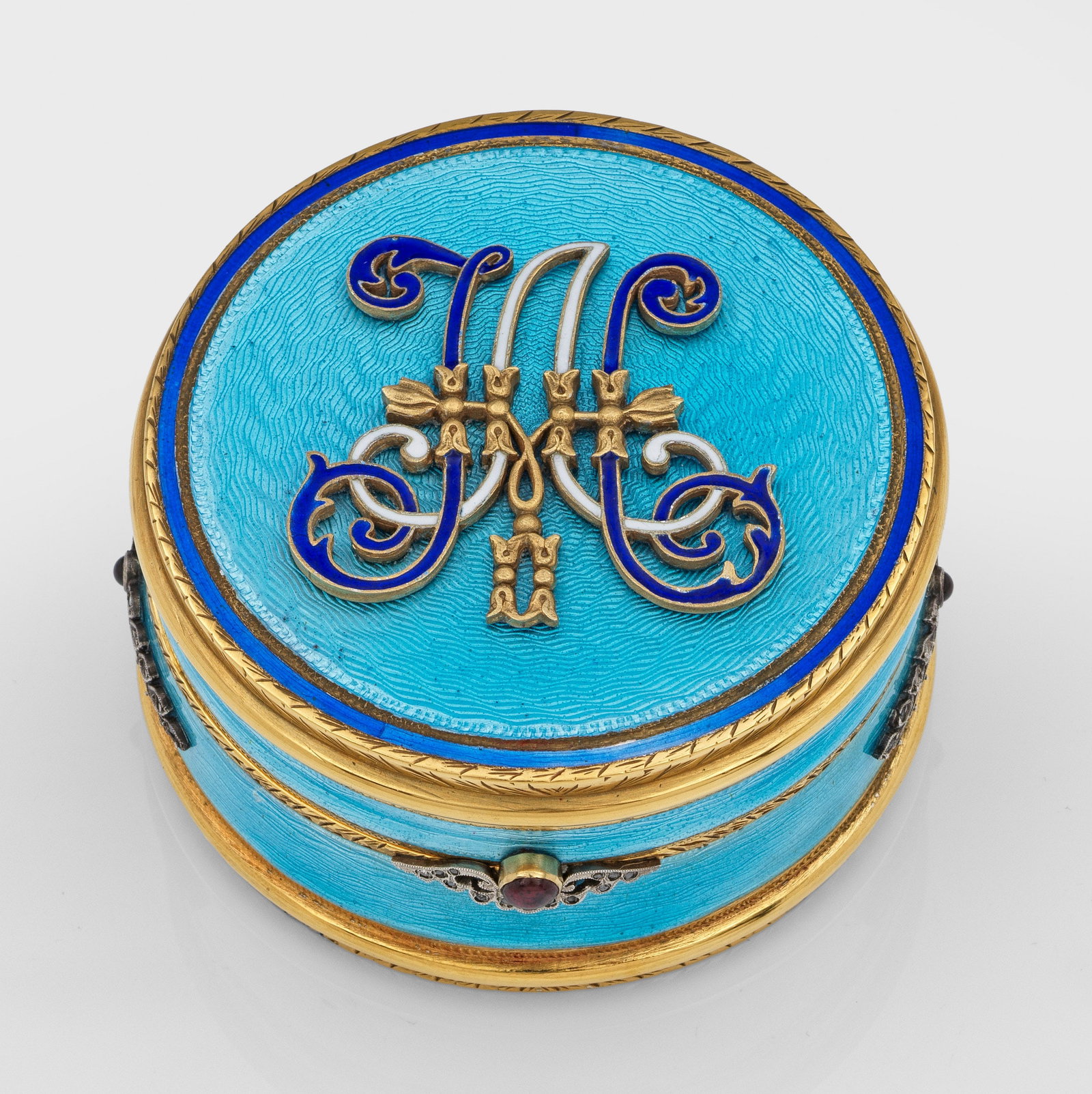 Runde Zierdose: im Stil von Karl Fabergé. Silber, vergoldet, teilw. emailliert. Flacher, zylindrischer Korpus. Auf flachem Scharnierdeckel mittig aufgelegtes blau und weiß emailliertes, ligiertes Monogramm über he