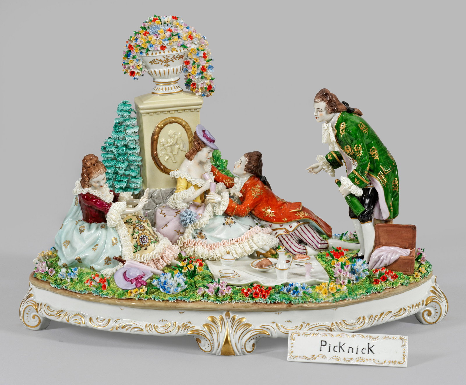 Große Figurengruppe "Picknick". Originaltitel: Ovaler, von Blattvoluten gezierter Sockel mit reliefplastischer Blütenwiese. Vor einem Picknick platziertes Liebespaar, sitzende galante Dame und Kavalier, ein Getränk kredenzend. Im Hintergund von 