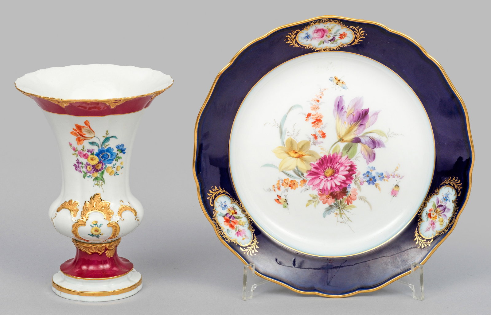 Zierteller und Kratervase mit Blumendekor: Runde, leicht vertiefte bzw. trichterförmig ausschwingende Form. Russisch roter Teilfond mit Goldbronze-Rocaillen und kobaltblauer Fahne. Blumengebinde in polychromer Malerei mit reicher Goldstaffage