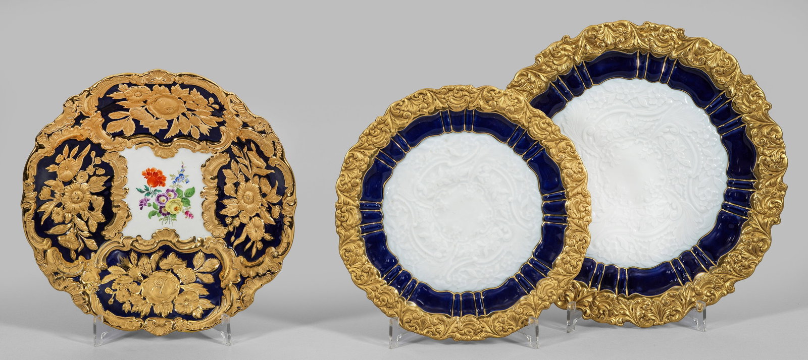 Prächtige Prunkschale und zwei Anbietplatten: 3-tlg.; Runde, leicht vertiefte bzw. flache Form mit passig geschweiftem Rand. Kobaltblauer Fond mit reliefierten Rocaillereserven und Blumenbuketts in sog. Goldbronze bzw. im Relief. Einmal Blumengeb