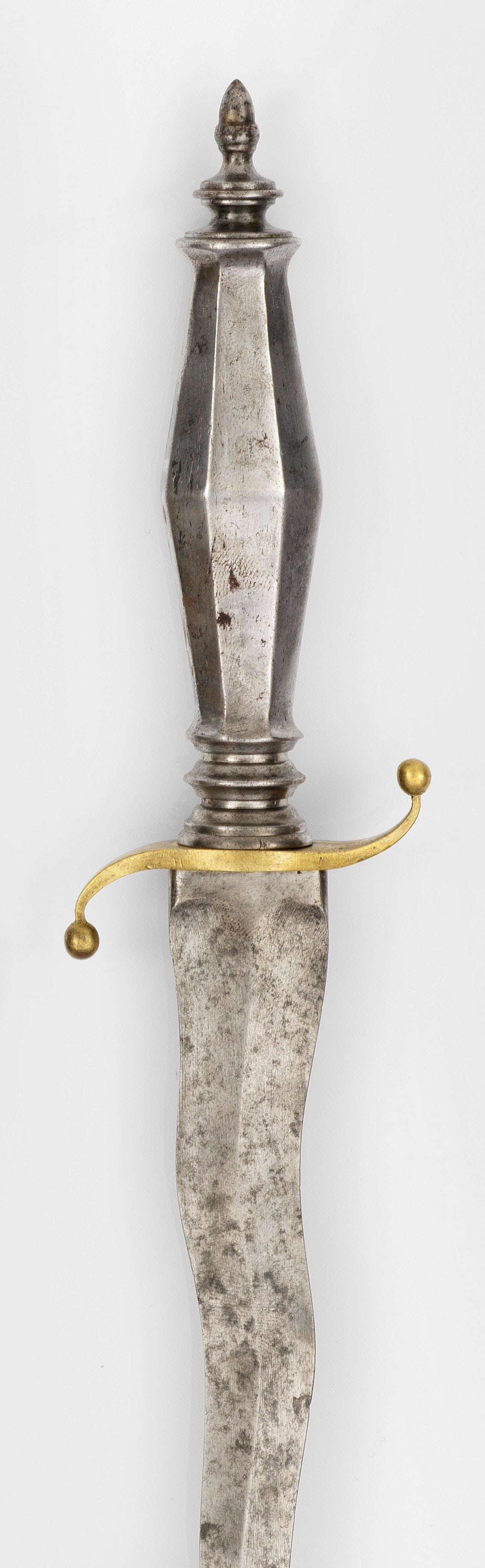 Historismus-Dolch mit geflammter Klinge: Eisen, Messing. Geflammte, beidseitig geschliffene Klinge, auf- und abwärts geschwungene, in Kugeln endende Parierstange aus Messing und achtfach facettierter, in Eichelknauf endender Griff. L. 24,5 
